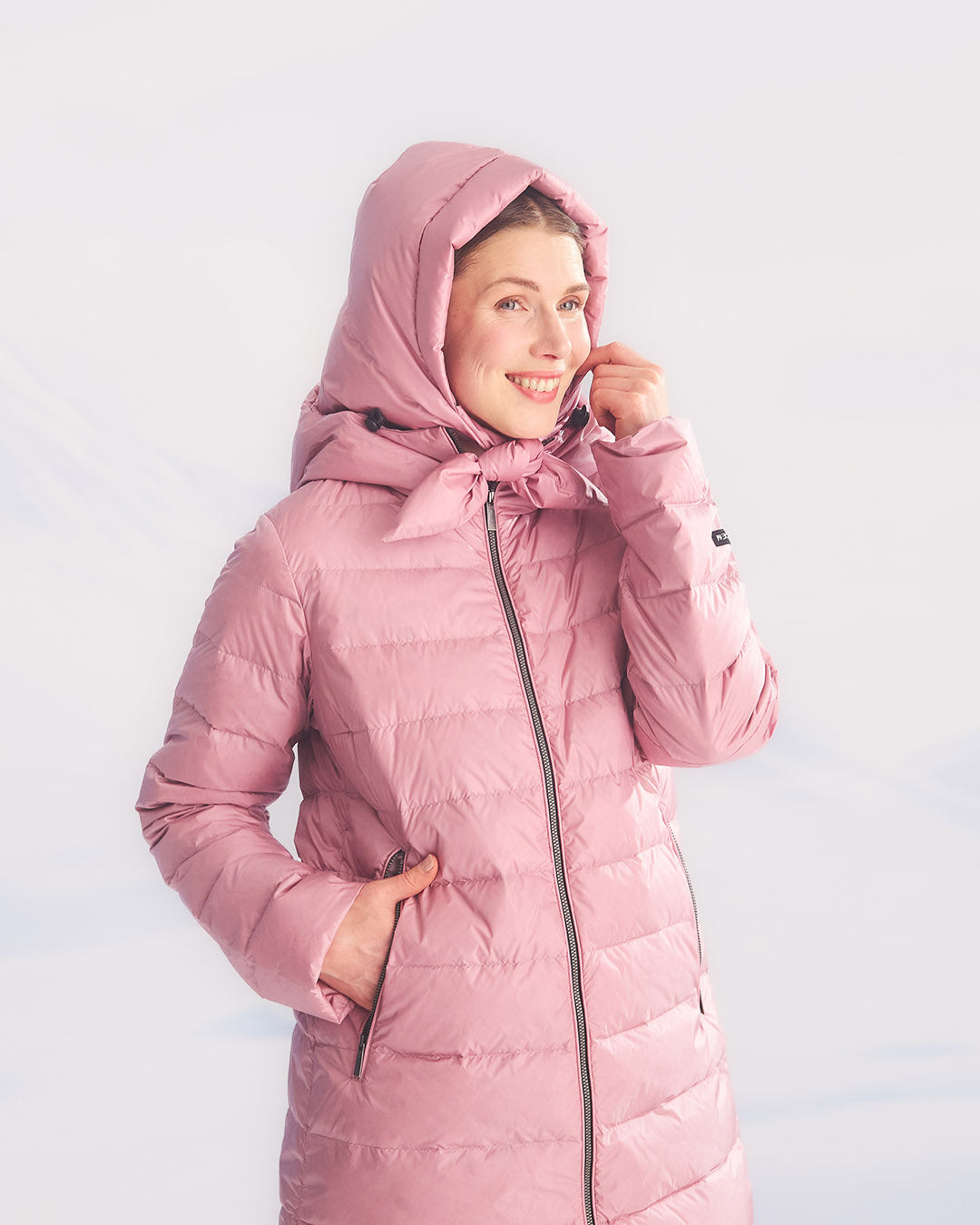 A smiling woman in a pink Ruut light down coat (color: Wildflower) with a detachable hood tied under the chin. Her hands are in the coat’s zippered pockets.