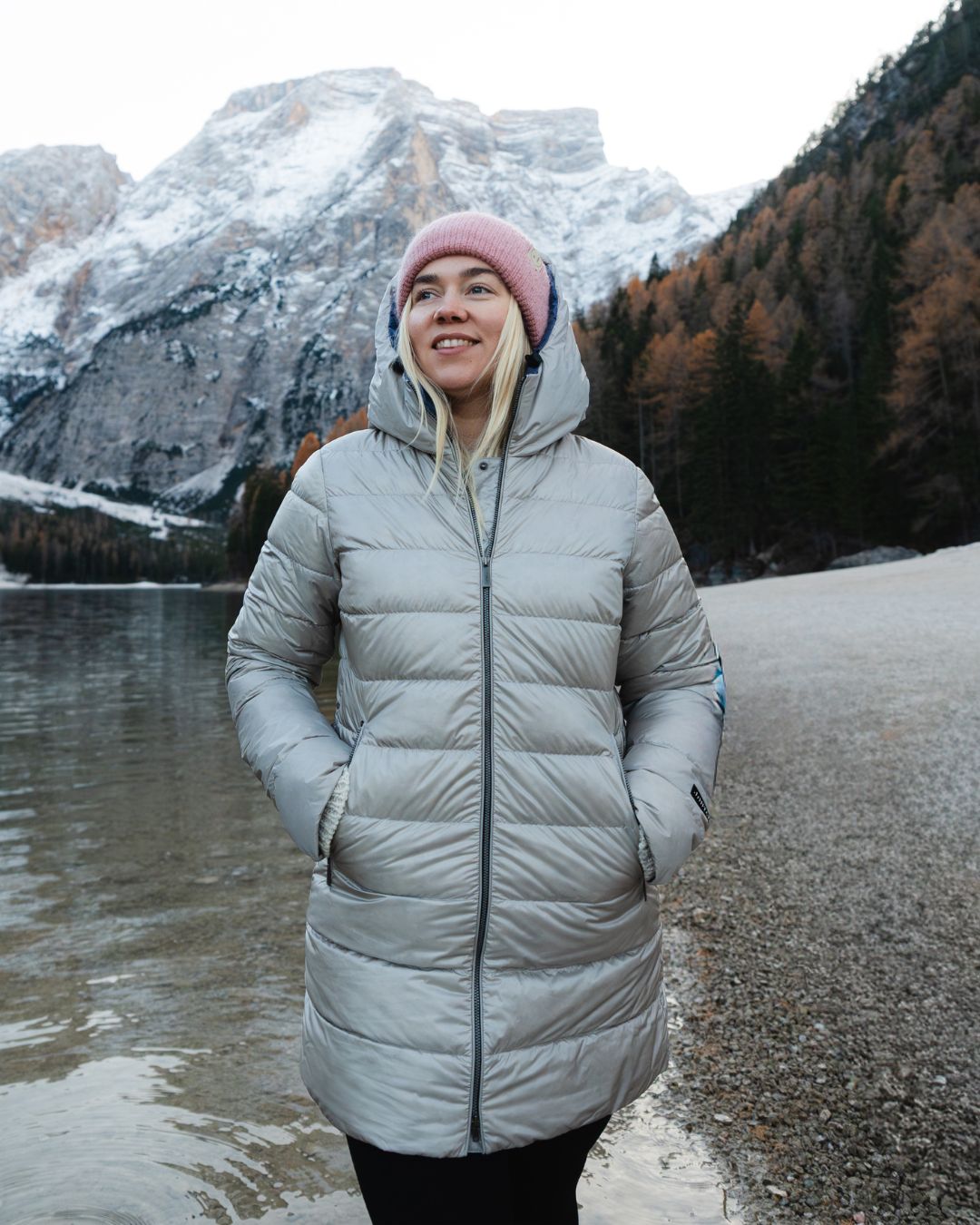 Person wearing a light gray (color_ Champagne) light down coat standing by a lake with mountains in the background