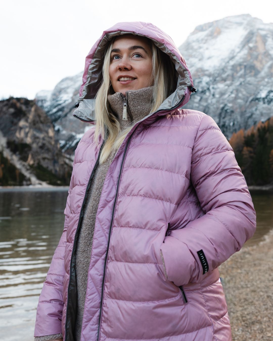 Person wearing a light pink (color: Wildflower) light down coat standing in front of a mountainous landscape.