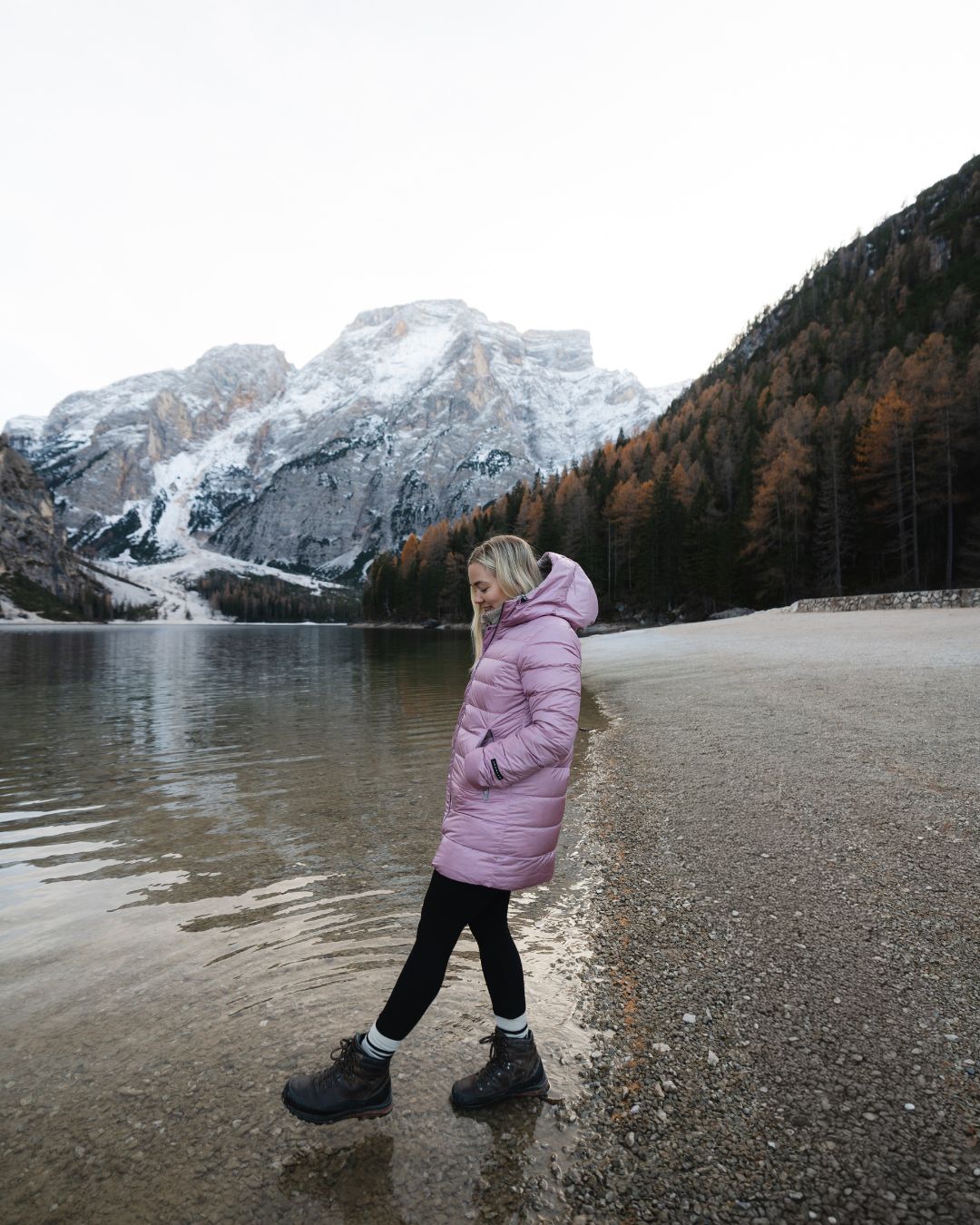 Person in a light pink coat walking near a lake with mountains in the background