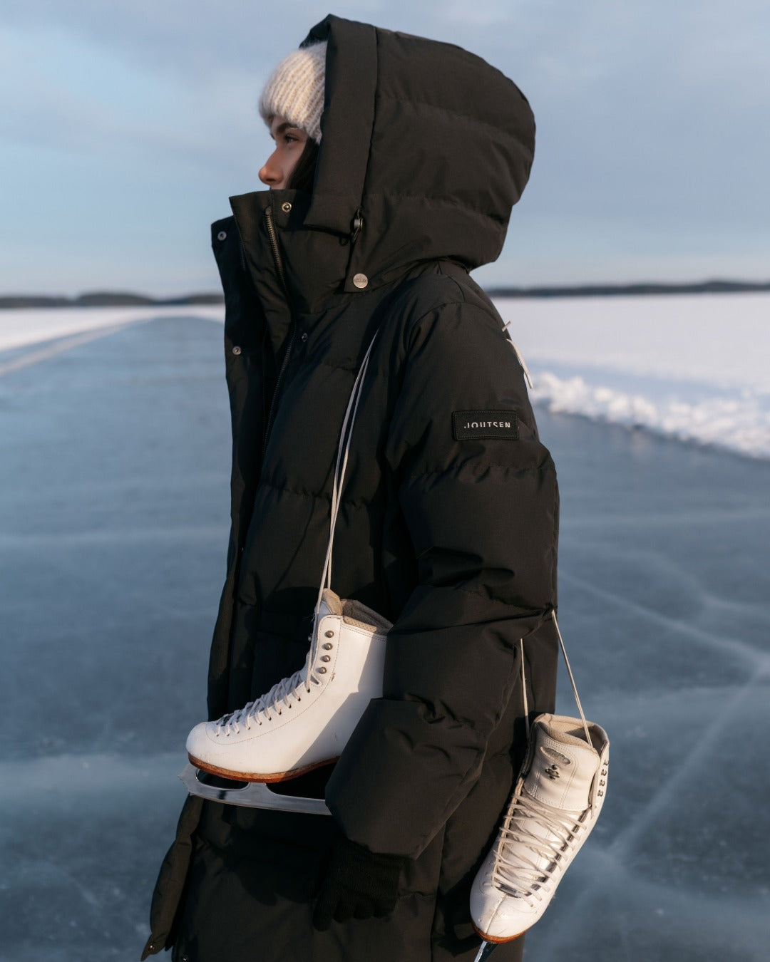Person in a black Siiri coat holding white ice skates on a frozen lake