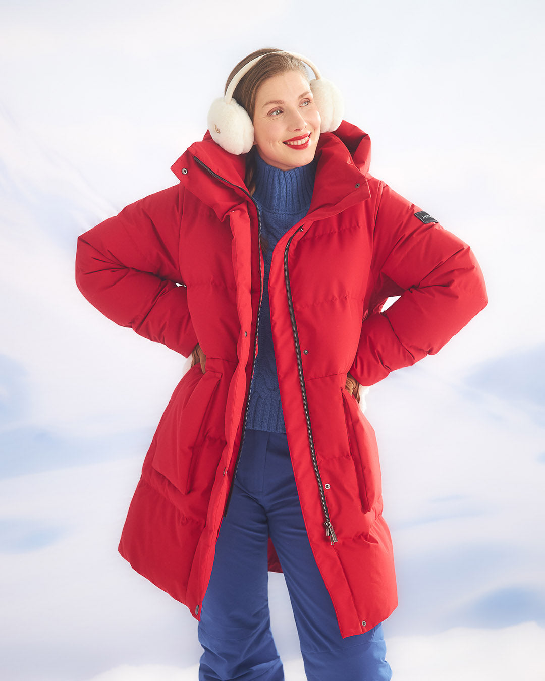 A woman is standing in a snowy landscape dressed in a red (color: chili pepper) Siiri down parka coat. The coat is long and equipped with a large hood as well as zipper and snap button closures. As accessories, she is wearing white earmuffs and blue insulated pants.