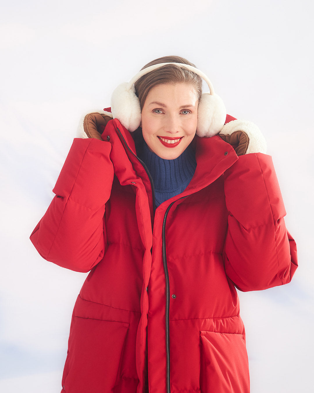 A woman is holding the hood of her red (color: chili pepper) Siiri down parka coat with both hands. The coat has large front pockets and a zipper. She is wearing large white earmuffs and a blue turtleneck sweater, with a light winter landscape in the background.
