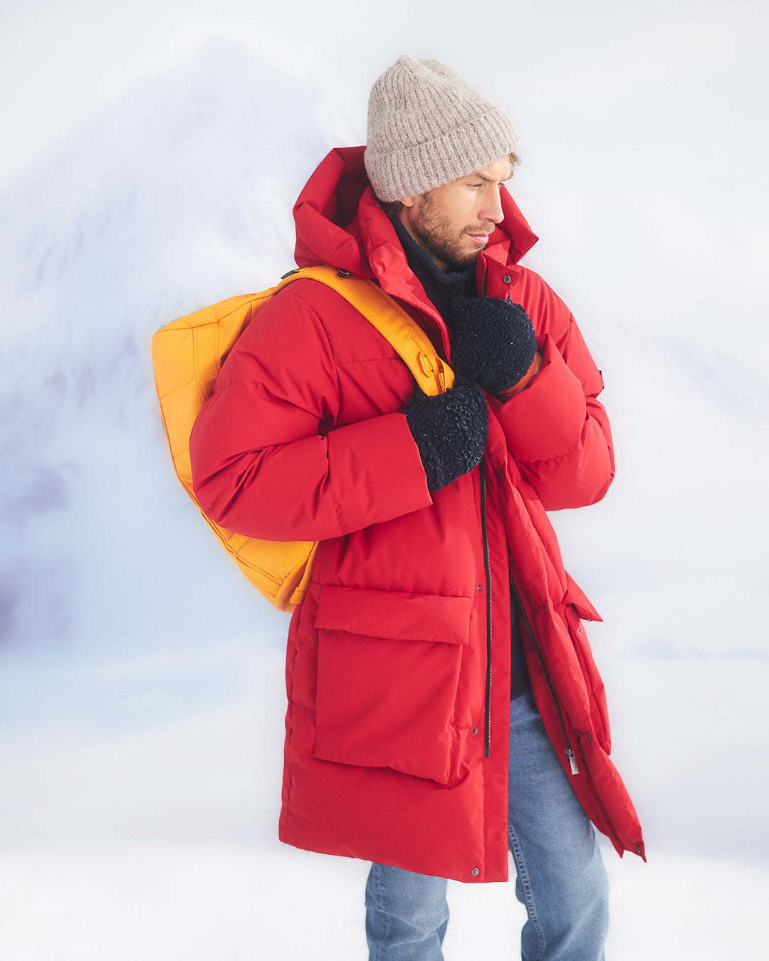 A man in a snowy landscape wearing a long, red (color: chili pepper) Tuisku down parka coat, a light grey beanie, blue jeans, and dark gloves. He is carrying a bright yellow backpack over his shoulder.