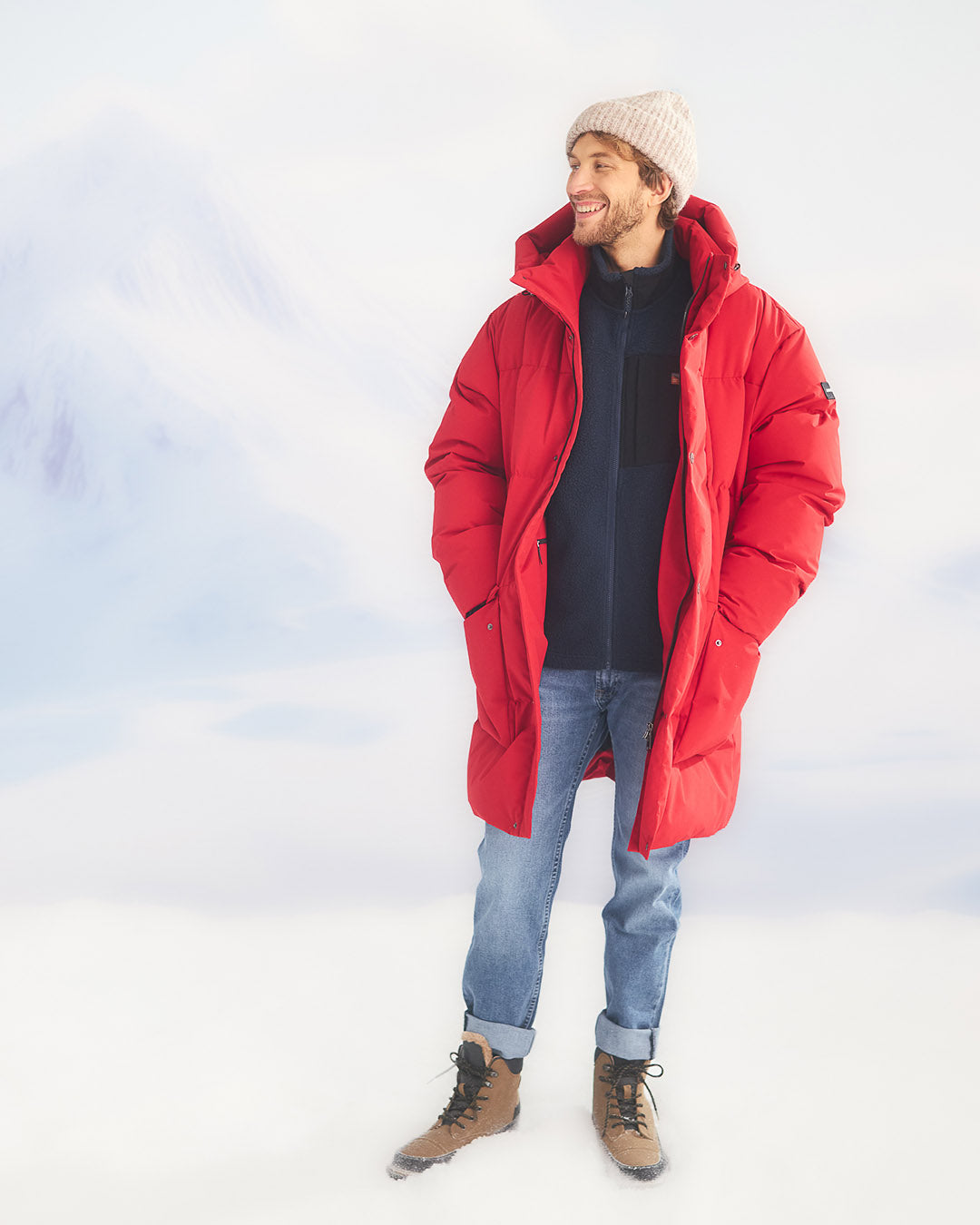 A man is standing in a snowy landscape wearing a long, red (color: chili pepper) Tuisku down parka coat left open at the front. Underneath, he has a dark blue fleece, blue jeans, a light grey beanie, and brown winter boots.