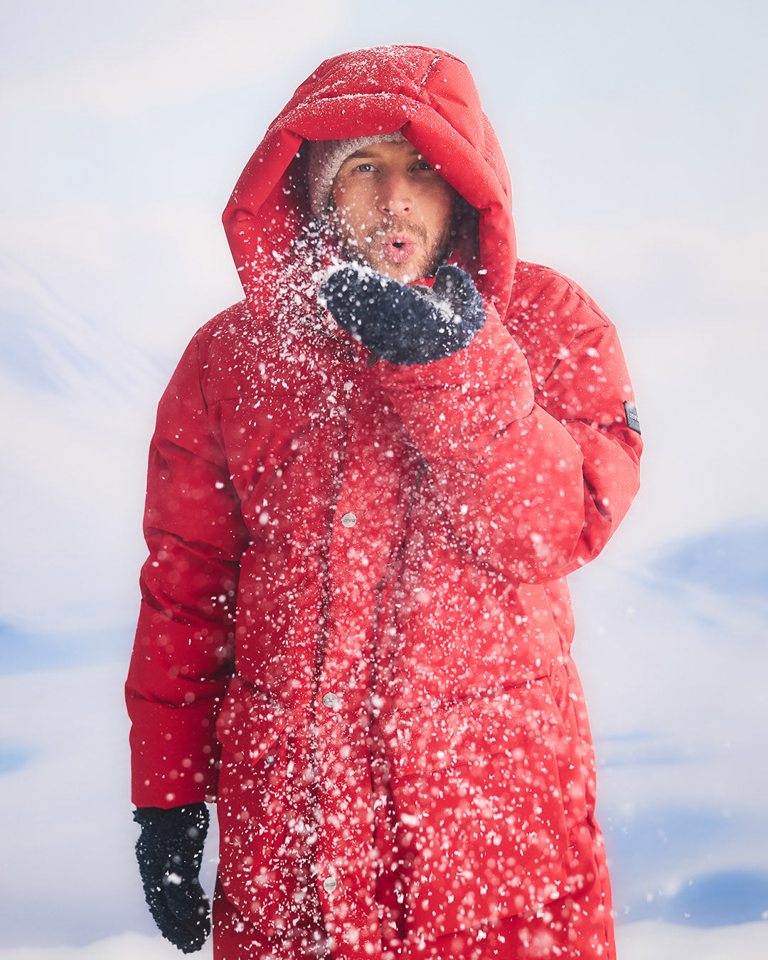 A man is standing in a snowy landscape, blowing snow from his hand so that the flakes drift into the air. He is wearing a long, red (color: chili pepper) Tuisku down parka coat with a large hood. On his hands, he has dark gloves.