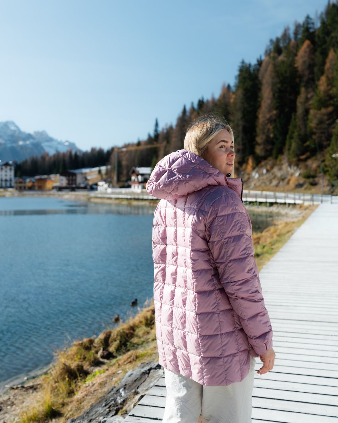 Woman in a pink coat standing by a lake with mountains in the background