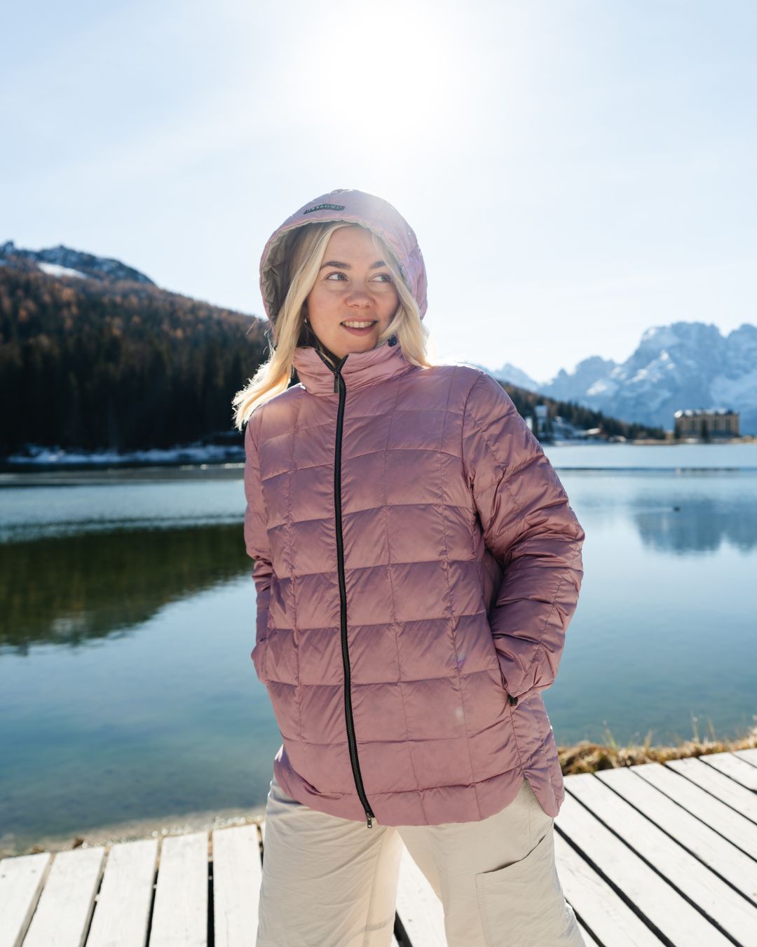 Person wearing a light pink down jacket standing by a lake with mountains in the background