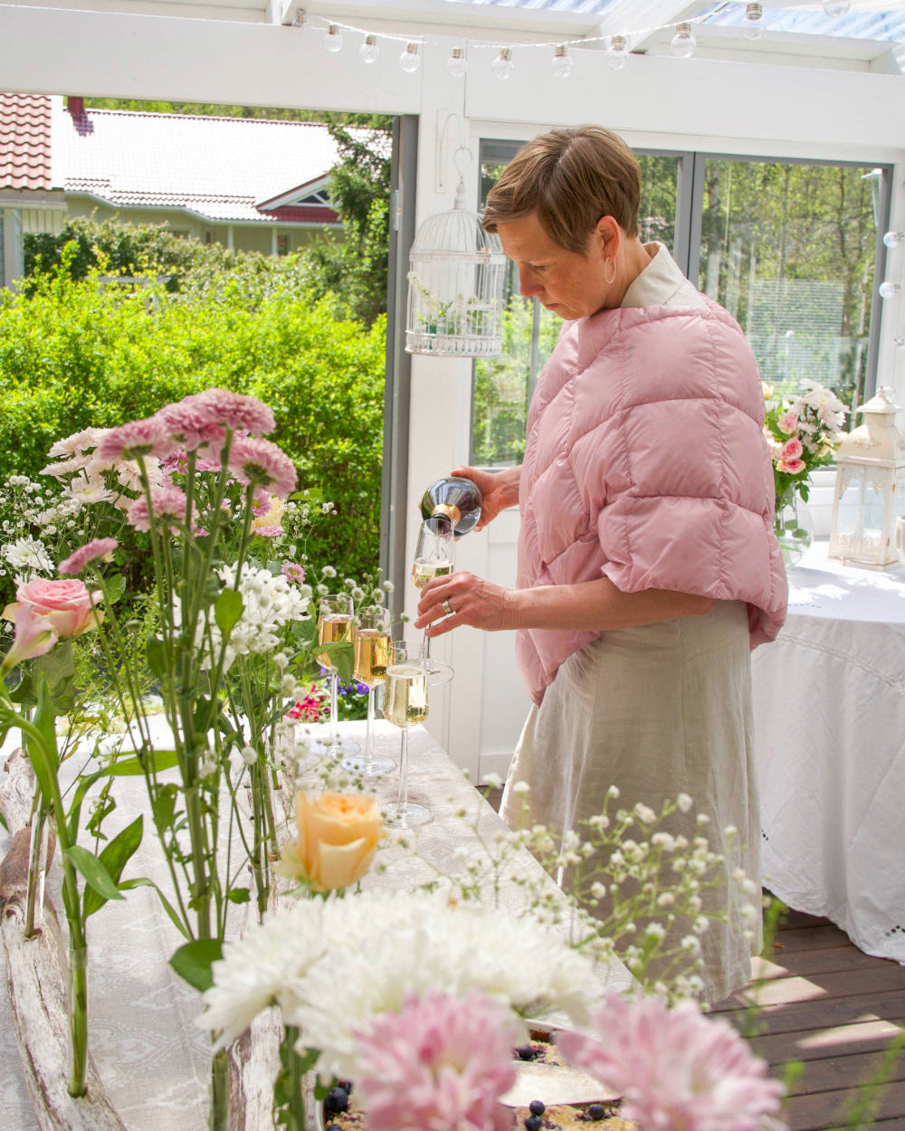 A woman stands on a bright veranda, pouring sparkling wine into glasses. She is wearing a light pink quilted Unelma down poncho (color: Rose) and a light linen dress. In the foreground, there is a long table decorated with an abundance of cut flowers, such as carnations, baby's breath, and roses. In the background, a lush garden is visible.