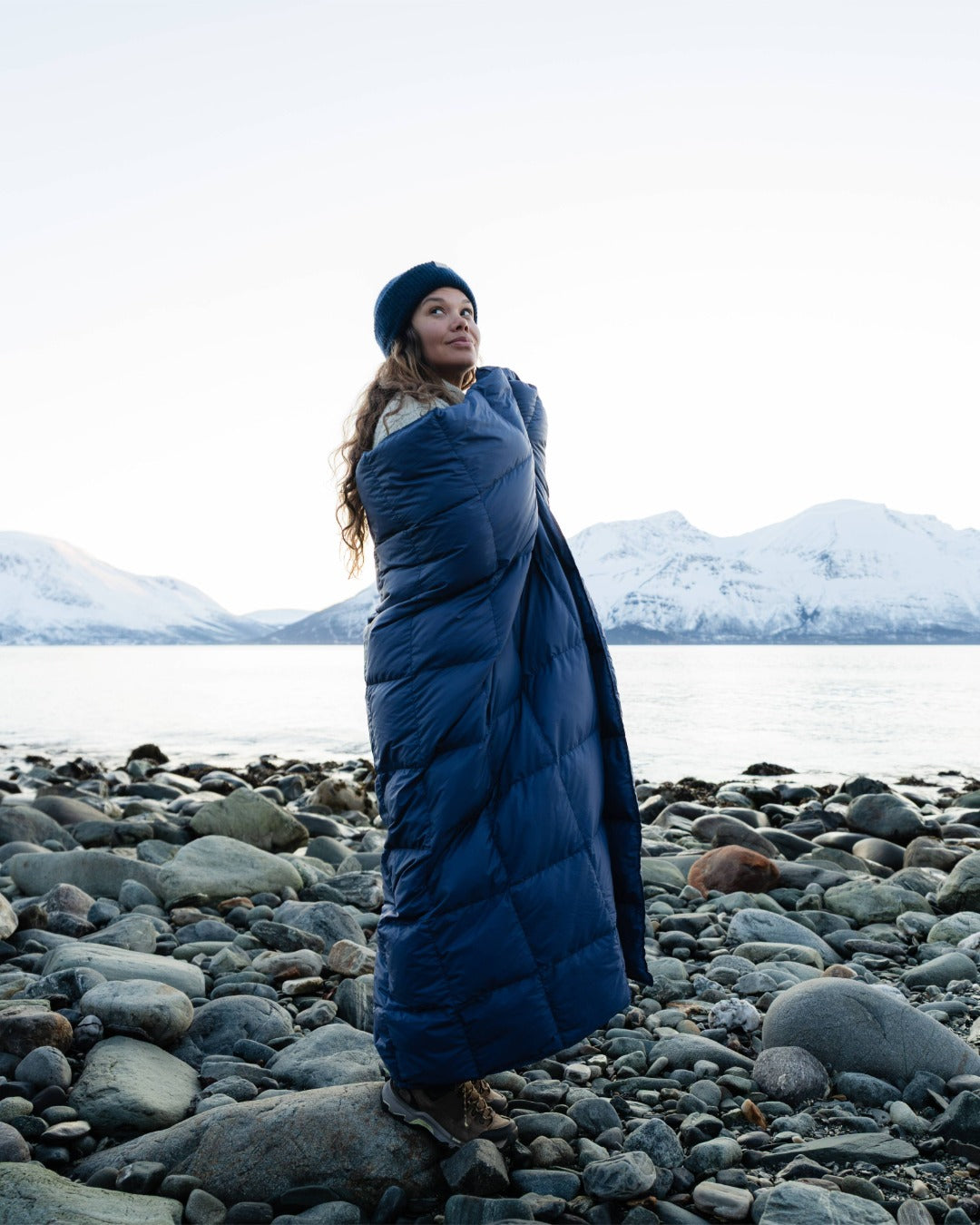 Person wrapped in a blue down blanket on a rocky beach with snowy mountains in the background
