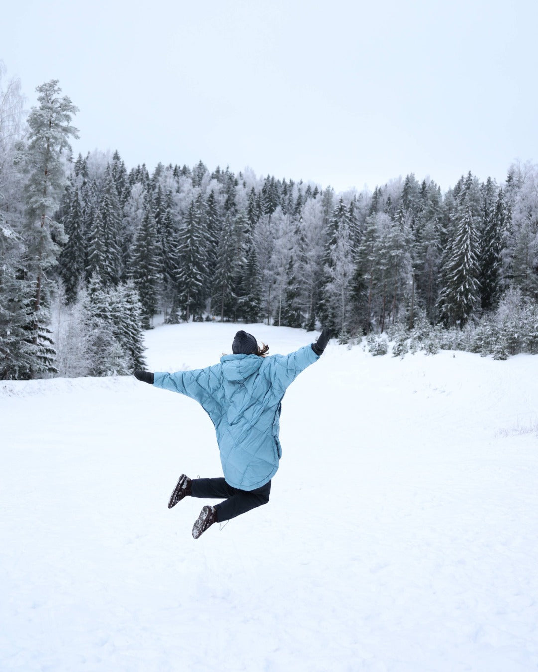 Person in a light blue down anorak jumping in a snowy landscape with trees
