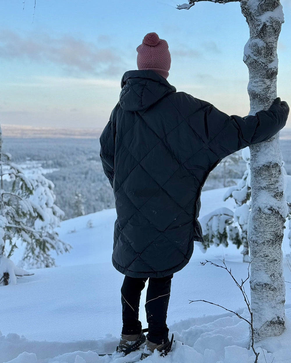 Person standing in snowy landscape wearing a charcoal black Velho down anorak with diamond quilting and a relaxed, oversized fit. The coat has a hood and a longer back hem, styled with a knitted beanie and winter boots.