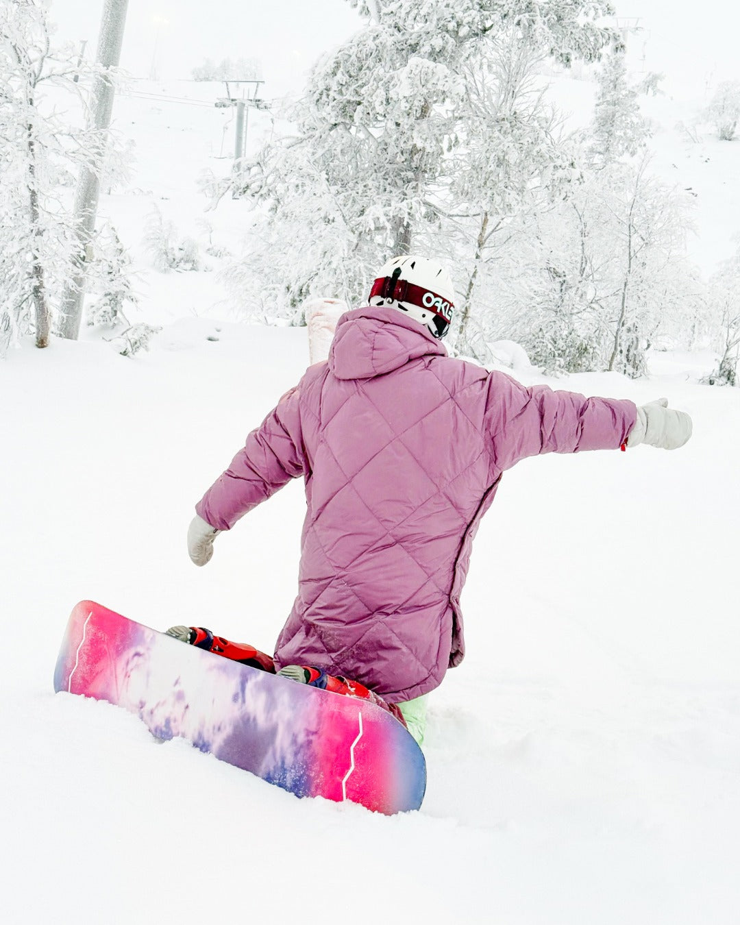 Person in a pink down anorak with a colorful snowboard, standing in a snowy landscape with trees.