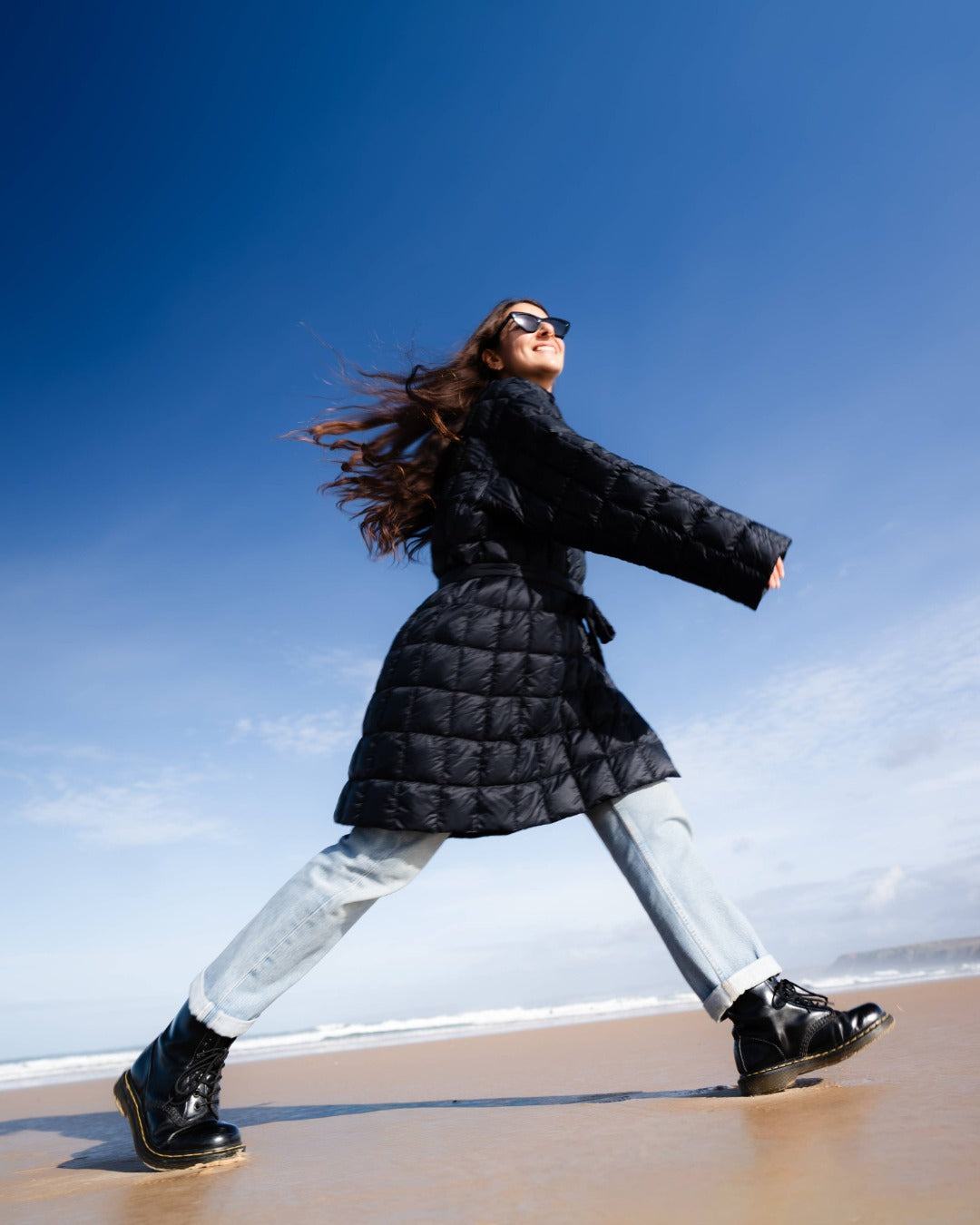 Woman in a black down jacket and white pants walking on a beach with a clear blue sky.