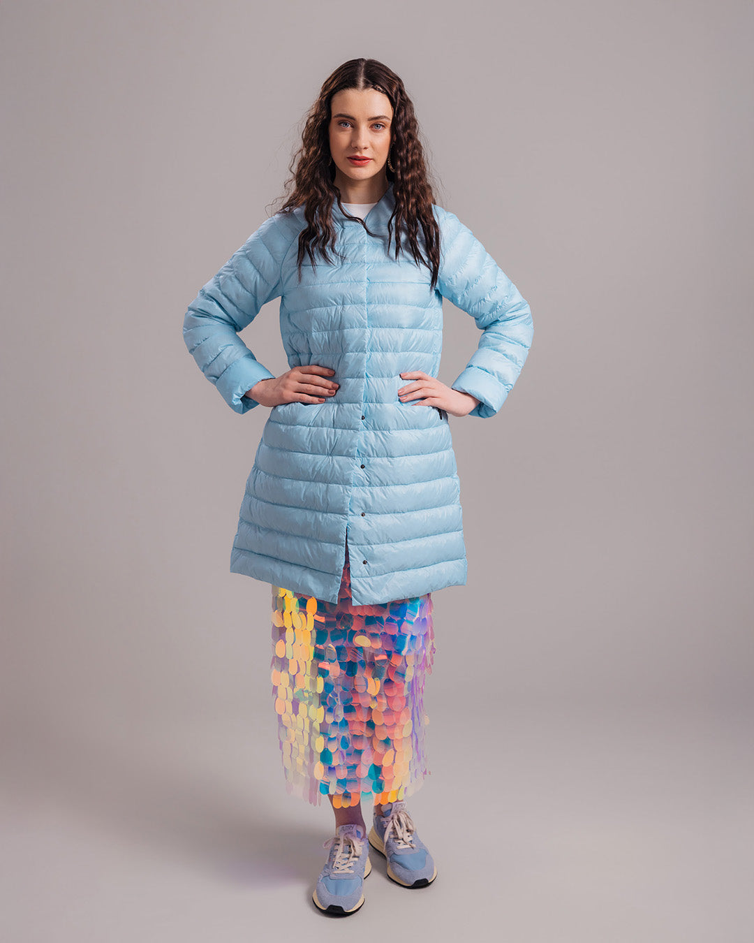 A woman stands with her hands on her waist, wearing a light blue, buttoned knee-length Vuokko light down jacket (color: Frozen candy), with a multicolored sequin skirt visible underneath. The outfit also includes blue sneakers.