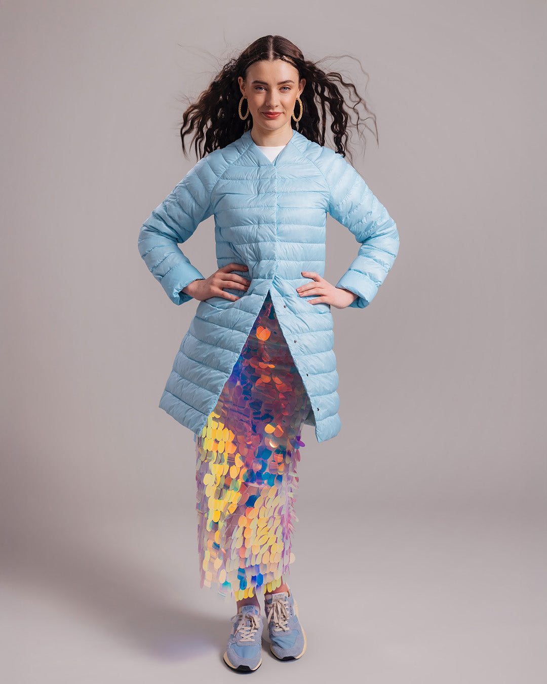 A woman dressed in a light blue, knee-length Vuokko light down jacket (color: Frozen candy), with a striking multicolored sequin skirt visible underneath. The outfit also includes blue sneakers and large earrings.