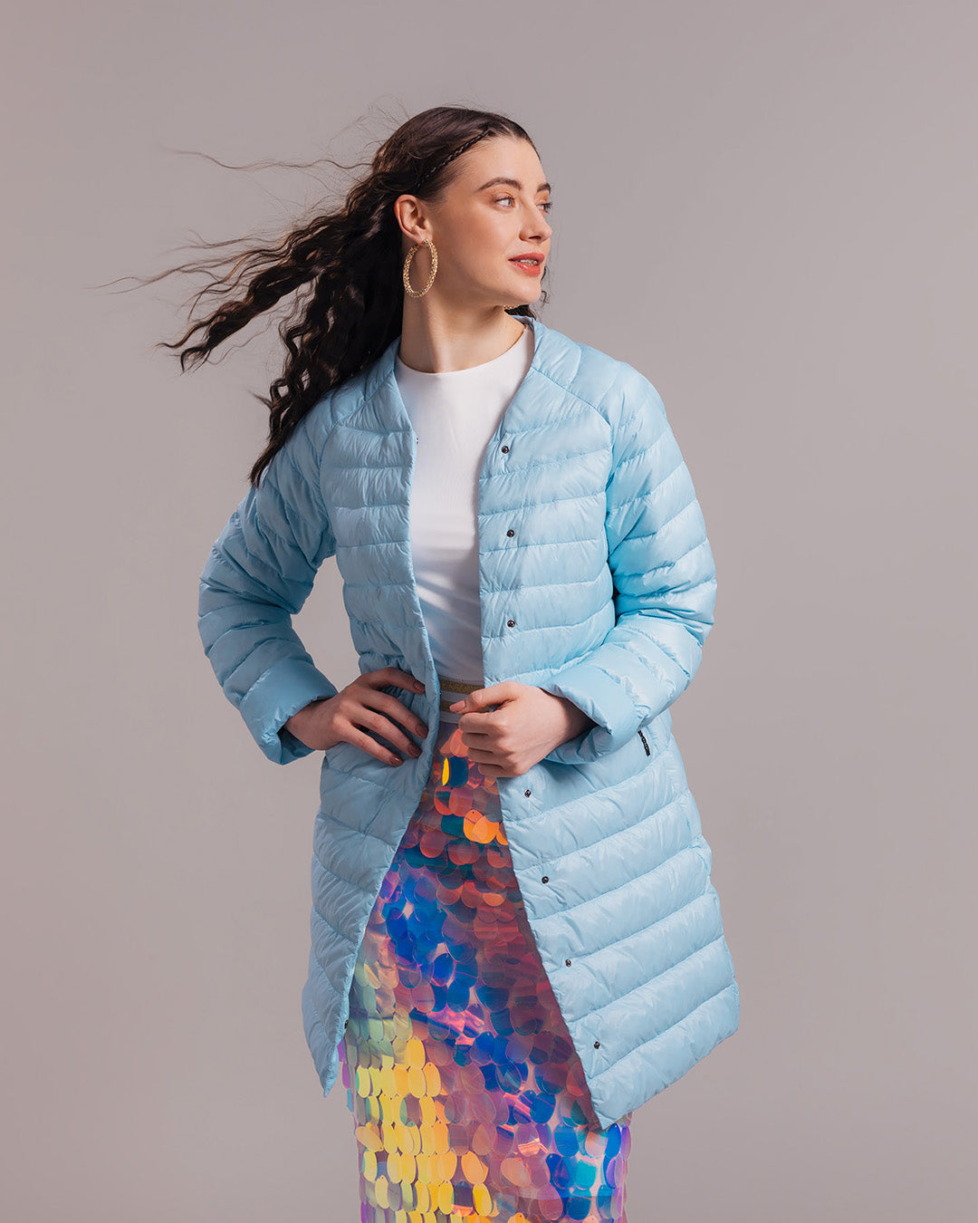 A woman dressed in a light blue, open, horizontally quilted, knee-length Vuokko light down jacket (color: Frozen candy), a white top, and a striking multicolored sequin skirt. The outfit also includes large hoop earrings.