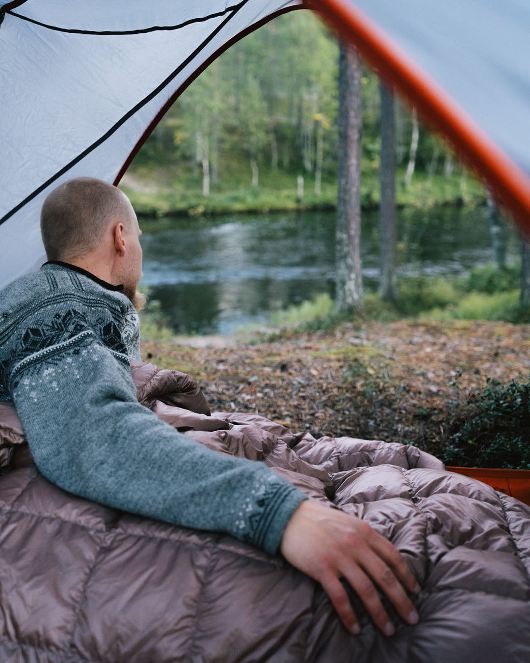A person wearing a blue wool sweater lies under a twilight grey Kulkuri down blanket inside a tent, looking towards the river. The tent opens to a view of a forested riverscape with mossy ground and trees along the riverbank.