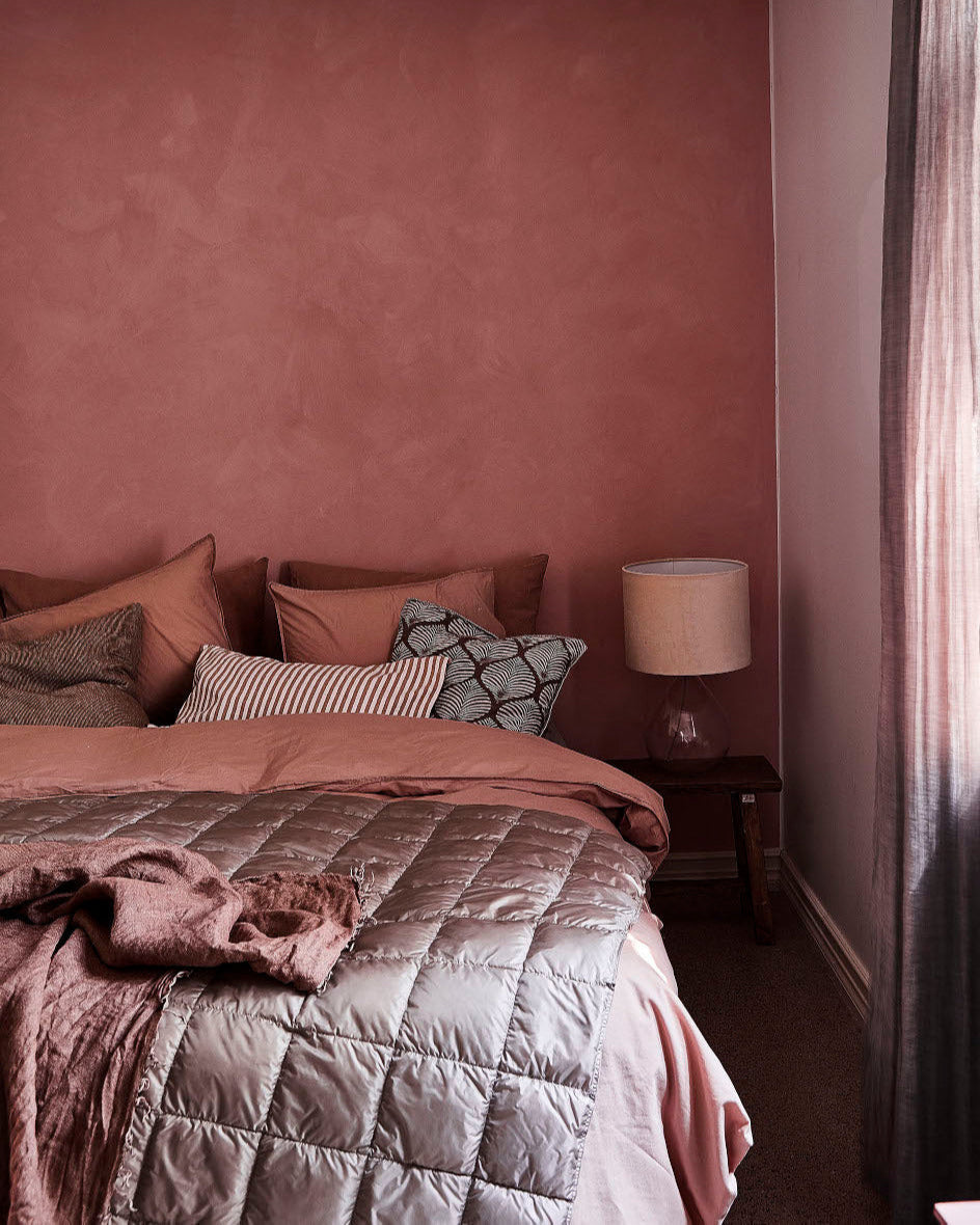 A bedroom with a bed made up in pink and brown toned bedding. On top of the bed lies a grey, quilted Kulkuri down blanket. In the background, there is a pink wall, a nightstand, and a table lamp. On the right edge, a sheer curtain and a window letting natural light into the room are visible.
