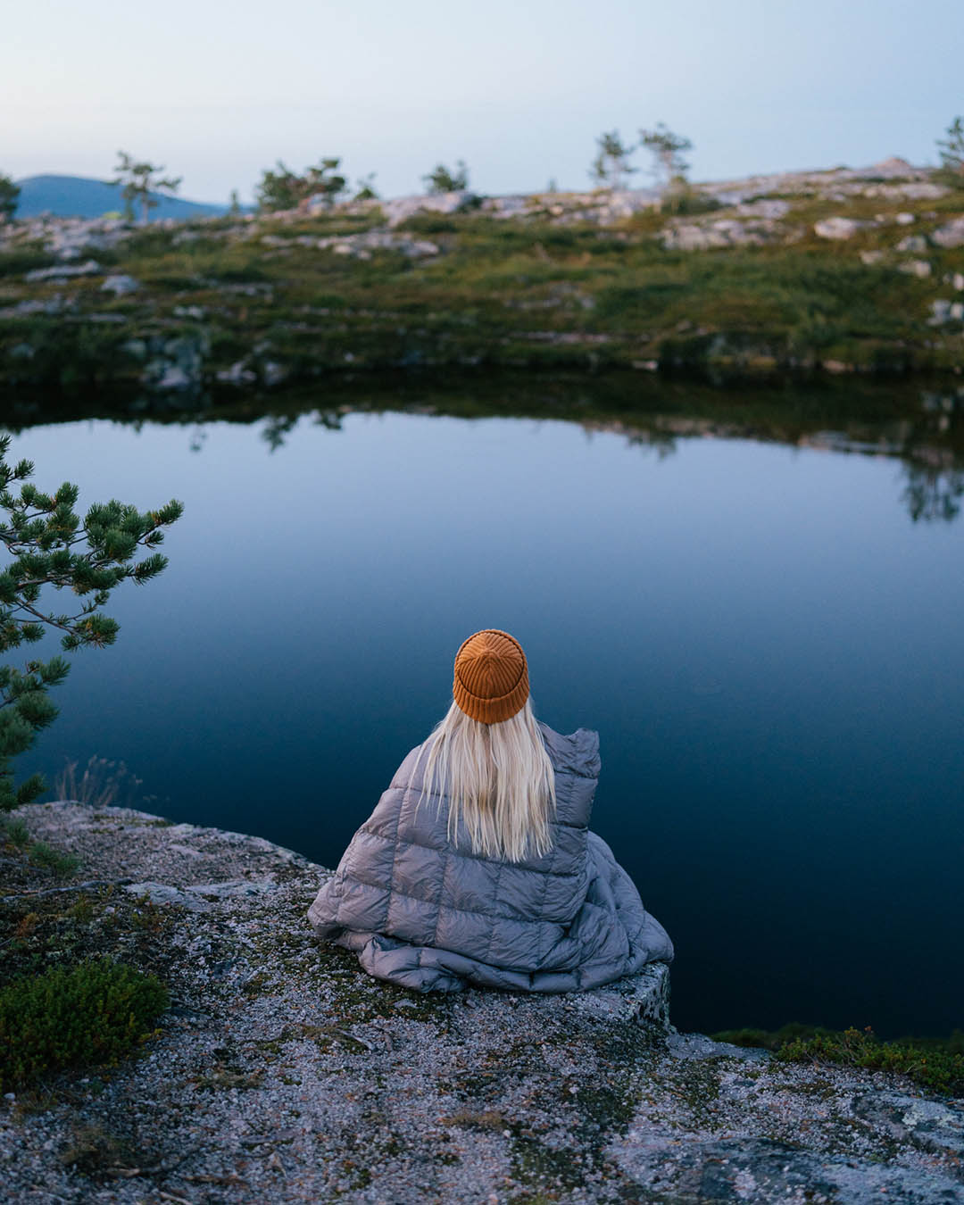A person is sitting with their back wrapped in a twilight grey Kulkuri down blanket by calm water in a rocky landscape. They are wearing an orange beanie and have long blonde hair. The water reflects the bluish sky, and in the background, tundra vegetation is visible.