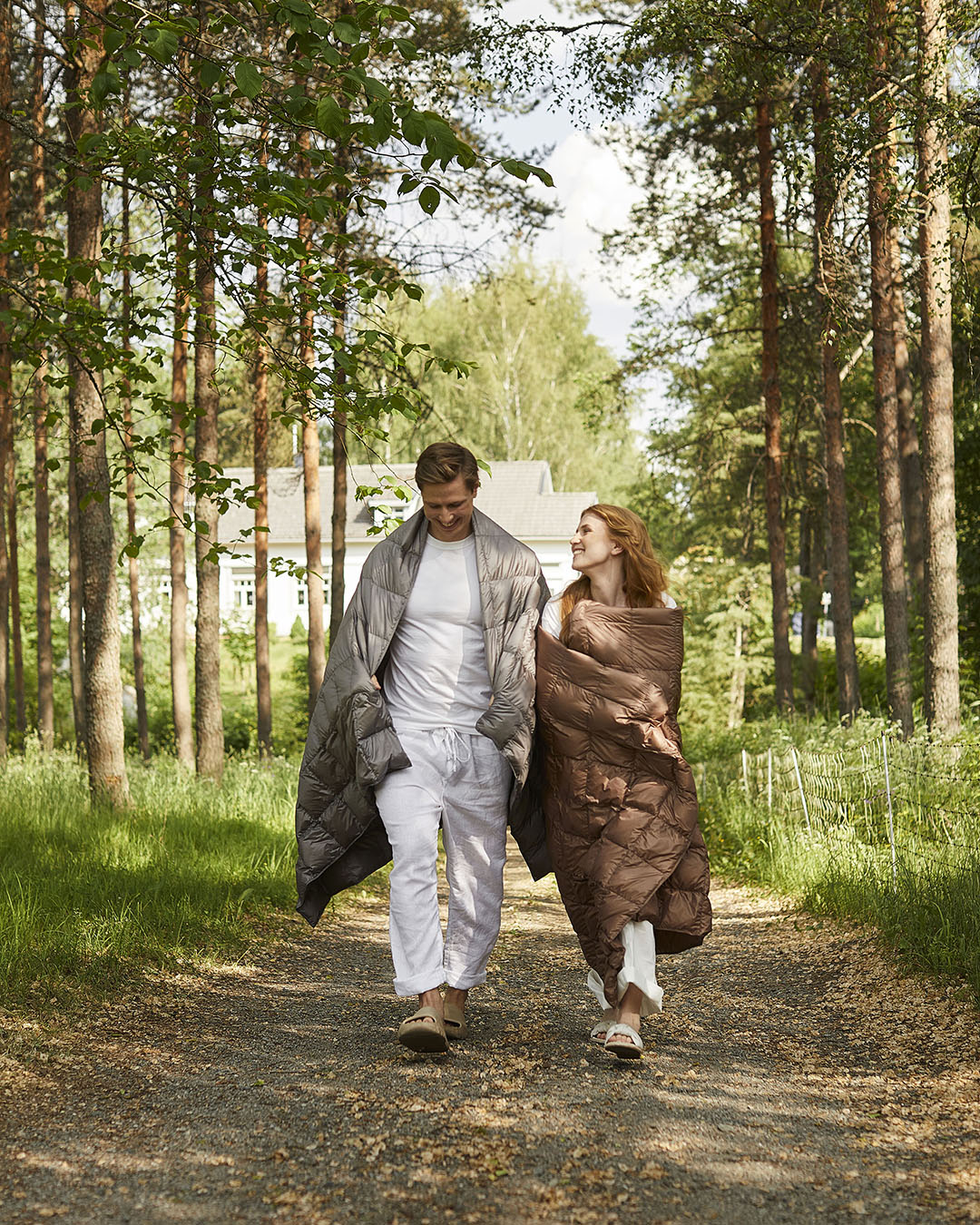 Two people are walking along a forested road, both wrapped in Kulkuri down blankets—one grey (twilight grey) and the other brown (color: hazelnut). They are smiling and looking at each other.