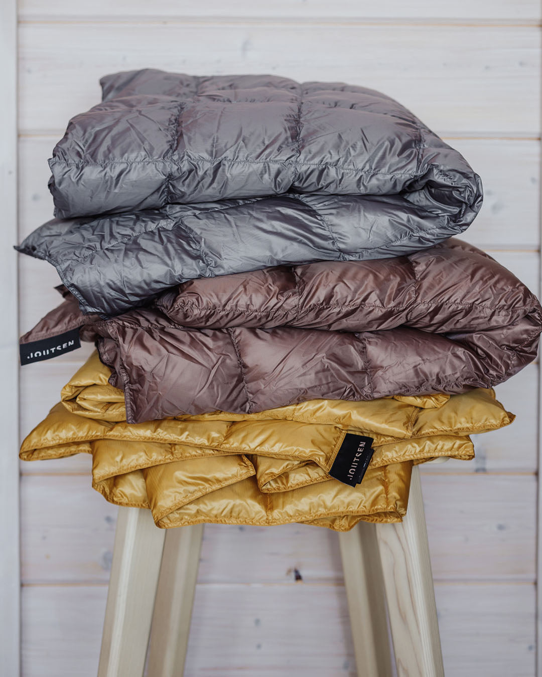 Three Kulkuri down blankets are neatly folded and stacked on a light wooden stool. The blankets are grey (color: twilight grey), brown (color: hazelnut), and yellow (color: Tuscan Sun), displaying the Joutsen brand label. In the background, there is a light horizontal paneled wall.