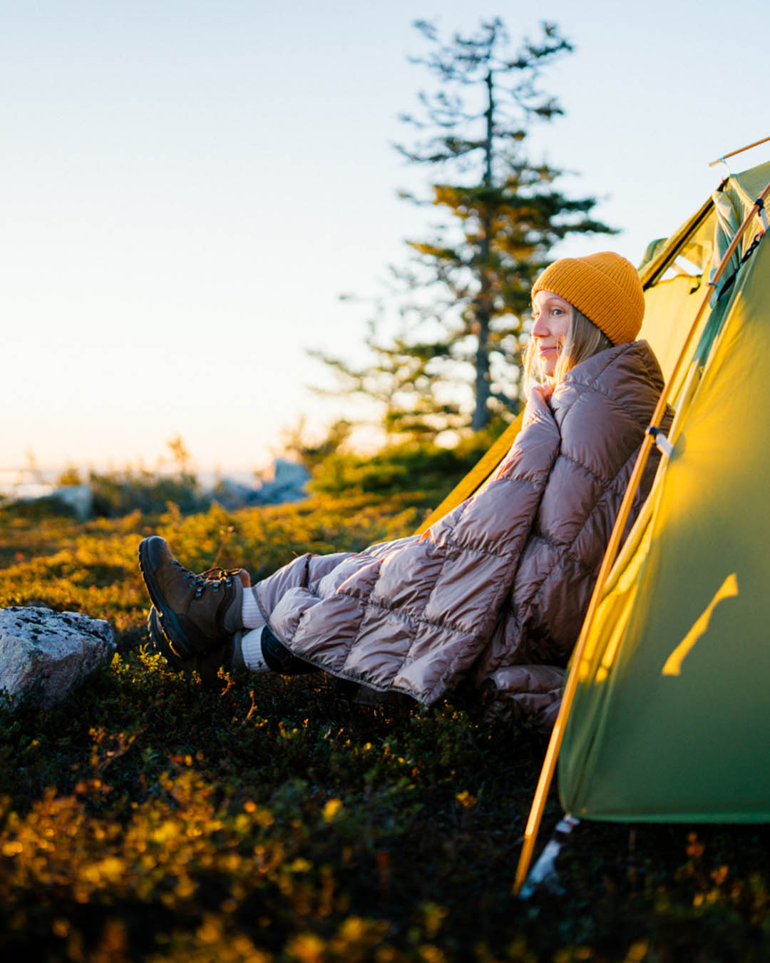 A person is sitting in front of a tent in nature, bathed in the morning sunlight, wrapped in a grey (color: twilight grey) Kulkuri down blanket. They are wearing a yellow beanie and hiking boots, looking toward the horizon.