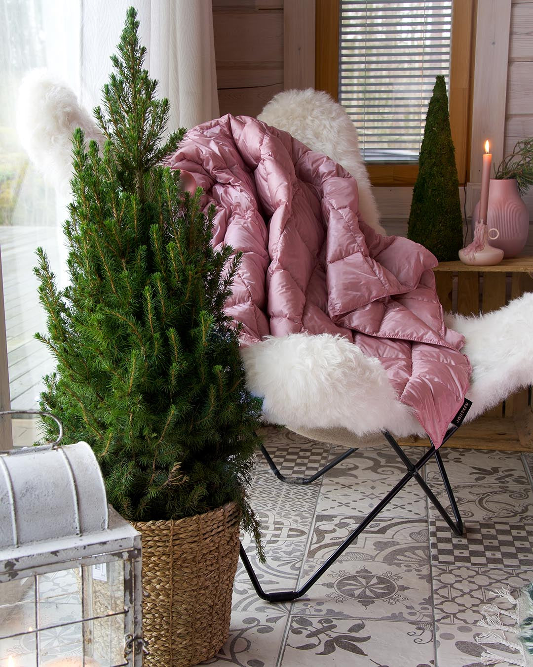 The light pink Kulkuri down throw blanket (color: rose) is draped over an armchair with a white sheepskin in a cozy room. In front, there is a small conifer tree in a woven basket, and in the background, a candle, decorations, and a large window are visible.