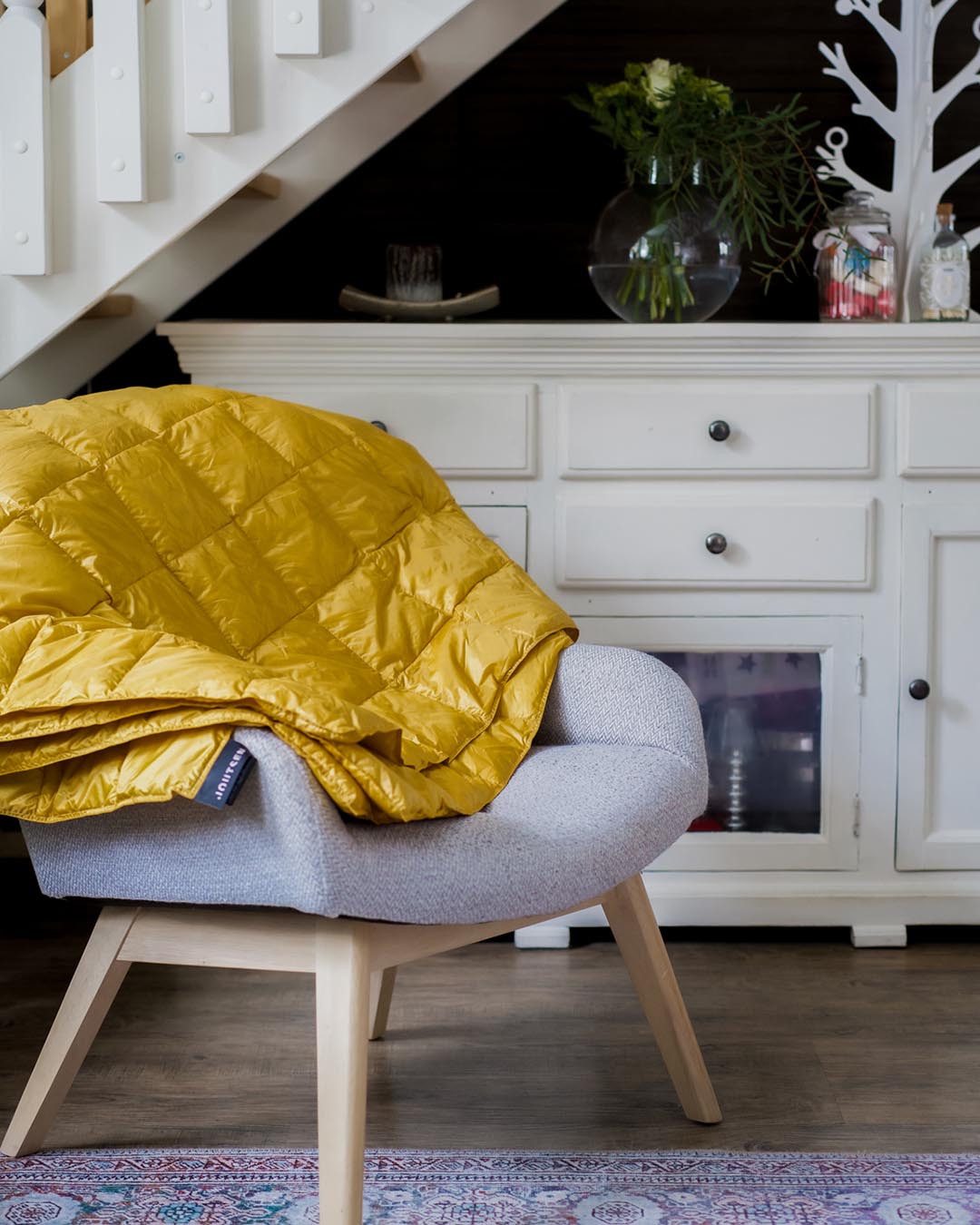 A yellow (color: Tuscan sun) Kulkuri down blanket is folded over the backrest of a light grey armchair in a cozy indoor setting. In the background, there is a white cabinet, a glass vase with green branches, and a decorative white wooden figurine.