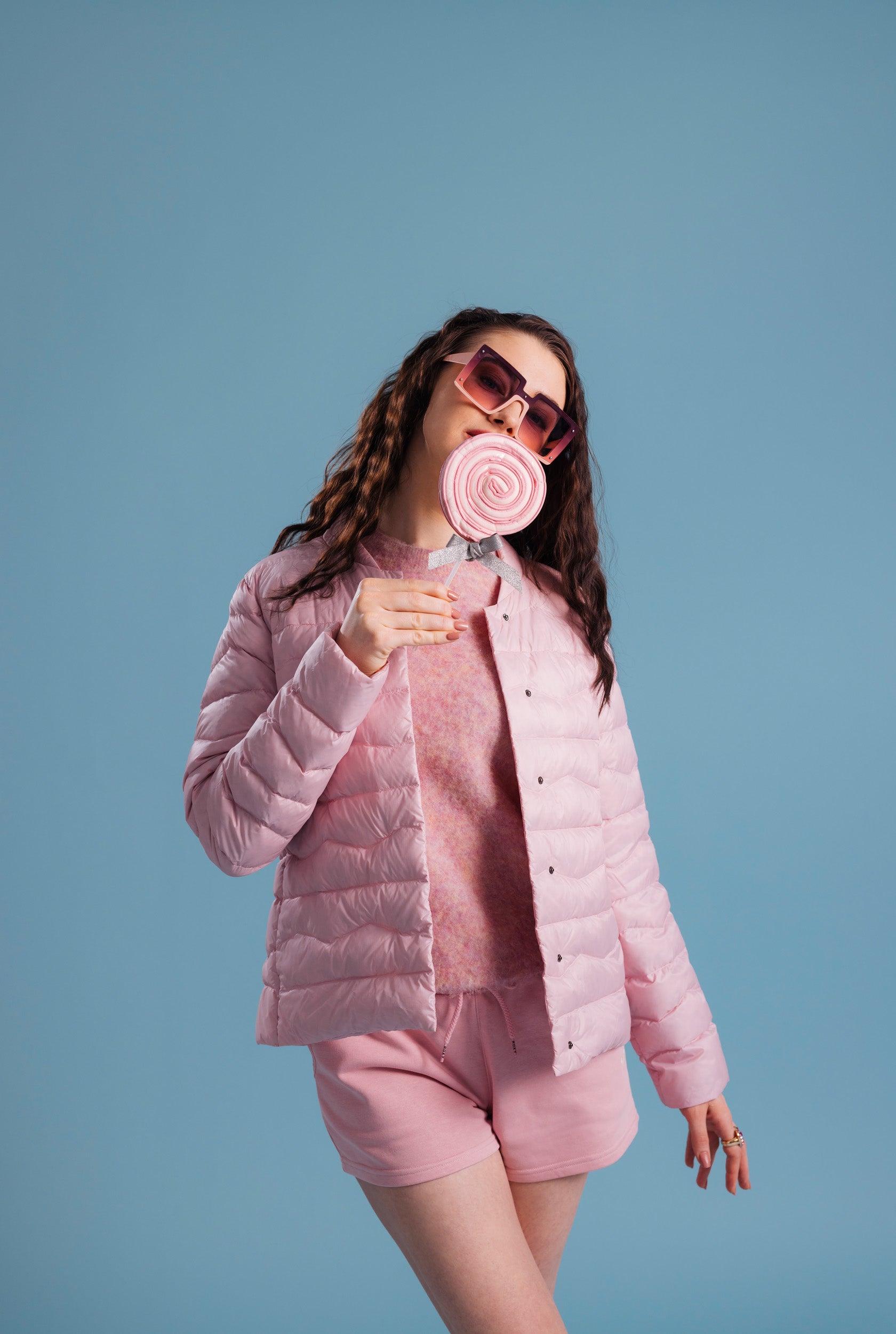 A woman dressed in a light pink Lootus light down jacket (color: strawberry mousse), a matching knit sweater, and shorts. The outfit is accessorized with oversized sunglasses and a round lollipop.