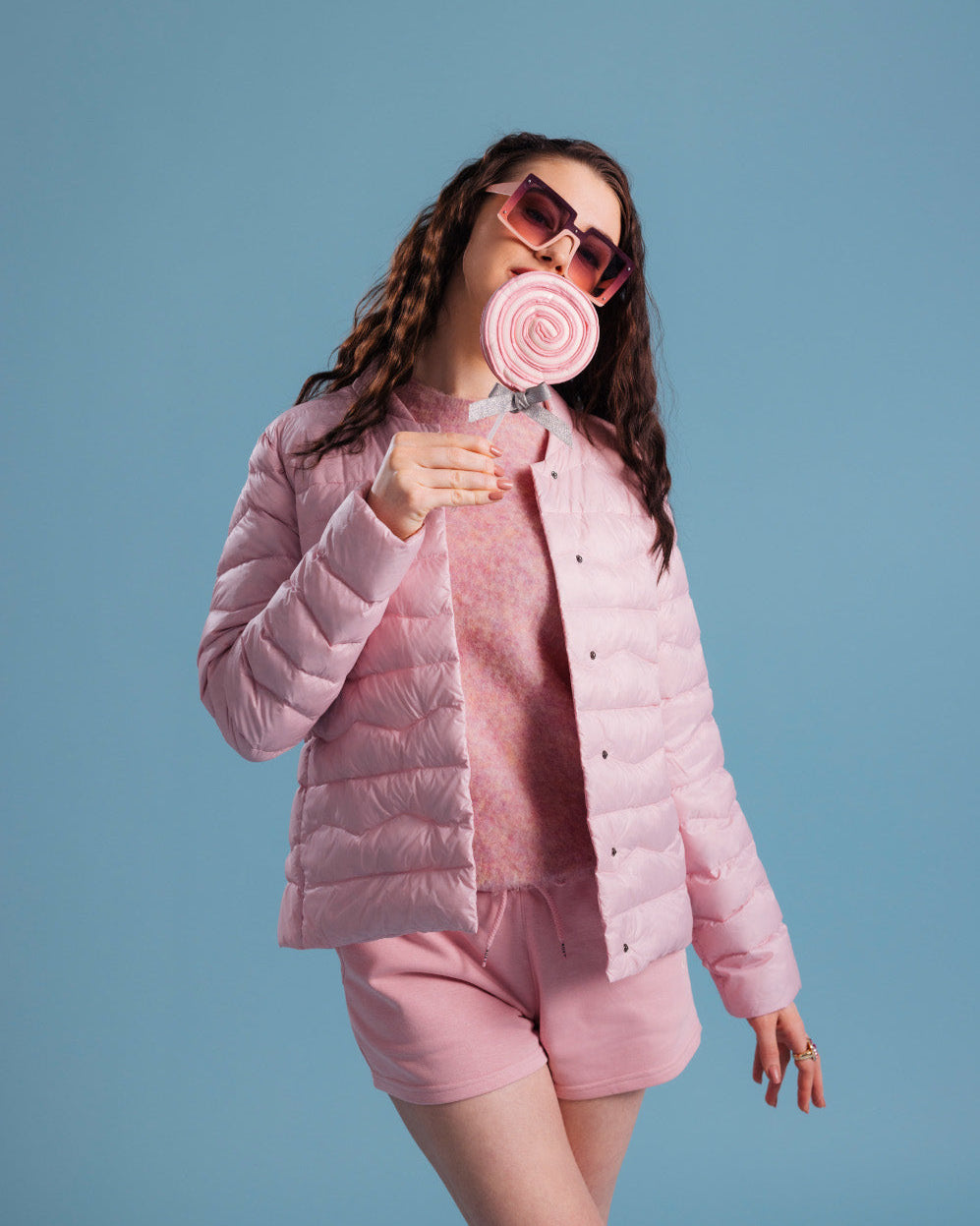 A woman wearing a pink Lootus light down jacket (color: strawberry mousse), a matching knit sweater and shorts. The outfit is complemented by oversized sunglasses and a round lollipop.