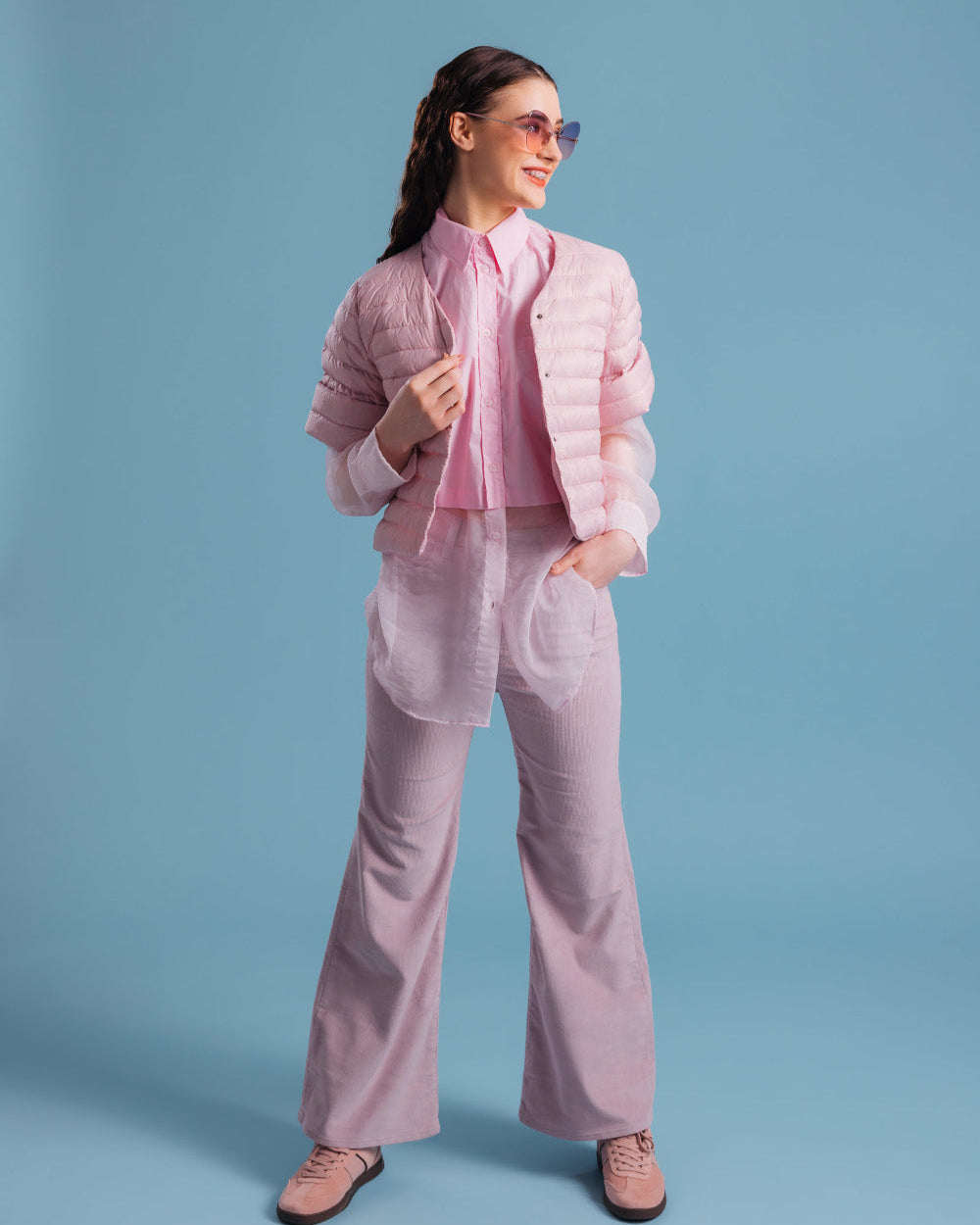 A woman wearing a light pink, horizontally quilted, short Lupiini light down jacket (color: strawberry mousse), styled over a long collared shirt and wide-legged trousers. The outfit is completed with sunglasses and pink shoes.