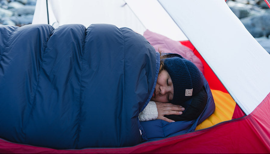 Person sleeping in a blue sleeping bag inside a red and white tent with a scenic background.