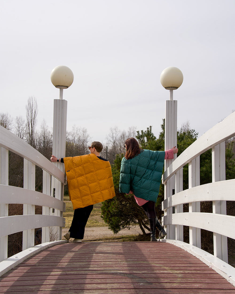 Two people are standing on an arched bridge with their backs to the camera, one wearing a yellow Otava down poncho (color: amber) and the other a green one (color: green paprika). In the background, a spring landscape with trees is visible.