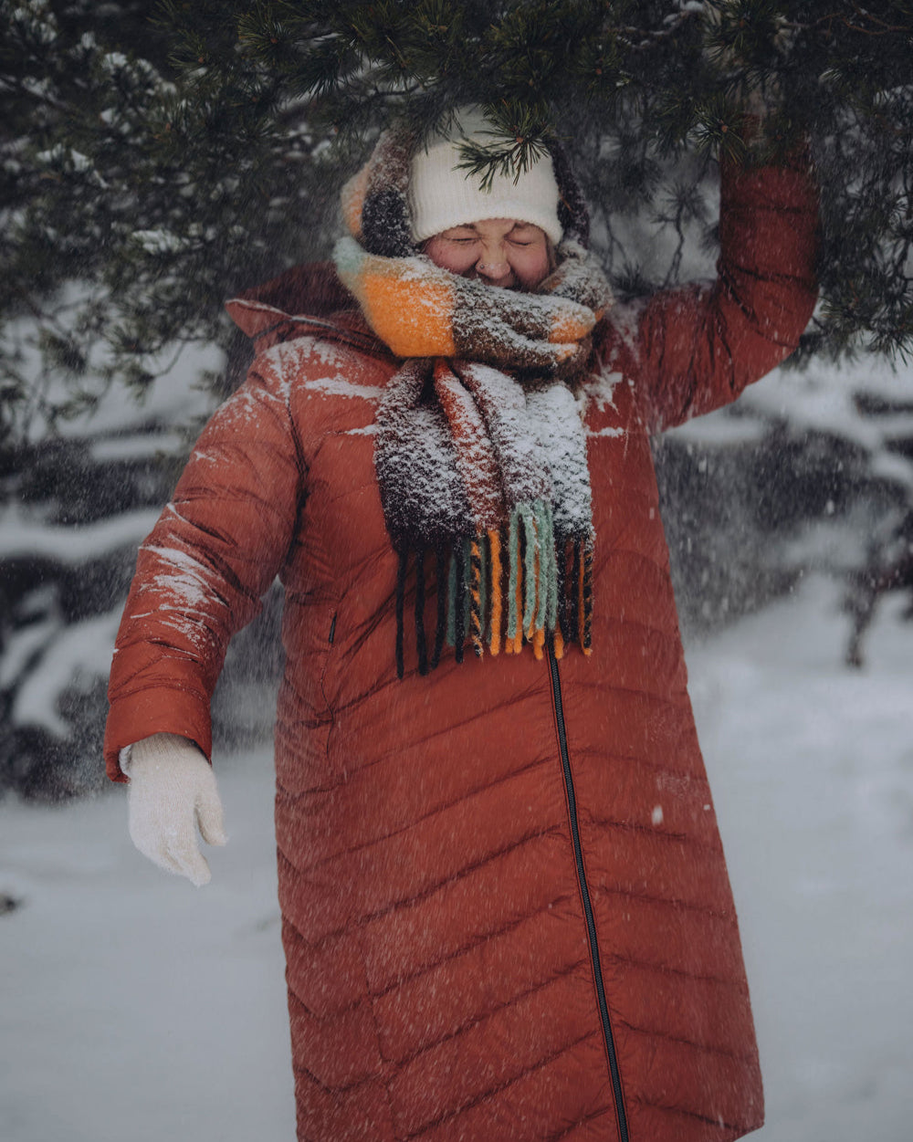 A woman is standing in a snowy forest wearing a muted orange Säde light down coat (color: rose gold), a white beanie, and a multicolored wool scarf. Snow is falling from a spruce branch onto her.