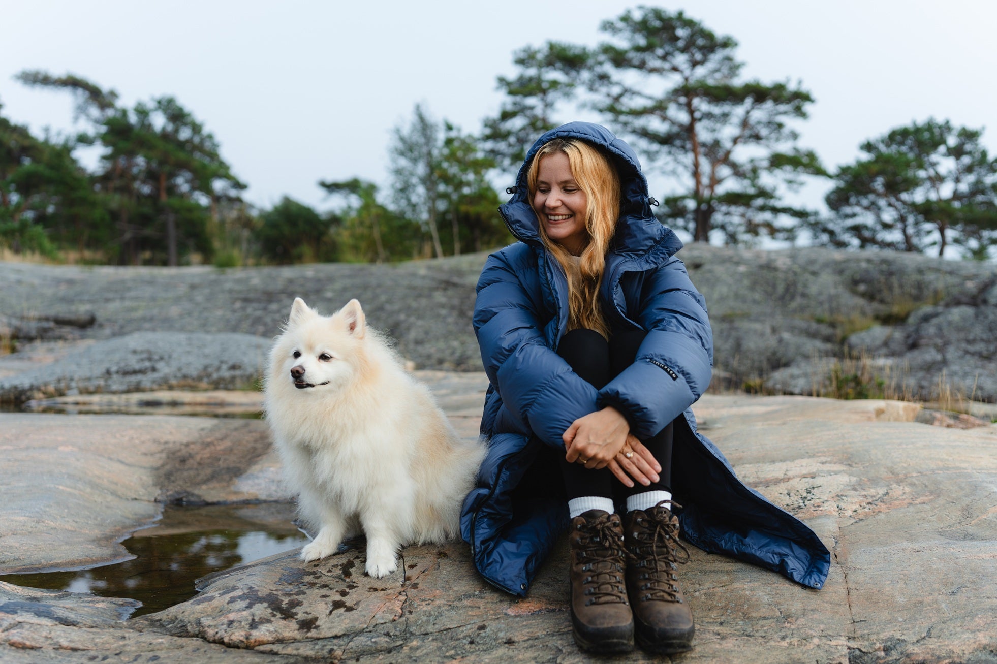 Woman in a blue down coat sitting on rocks with a white dog by her side, surrounded by trees.