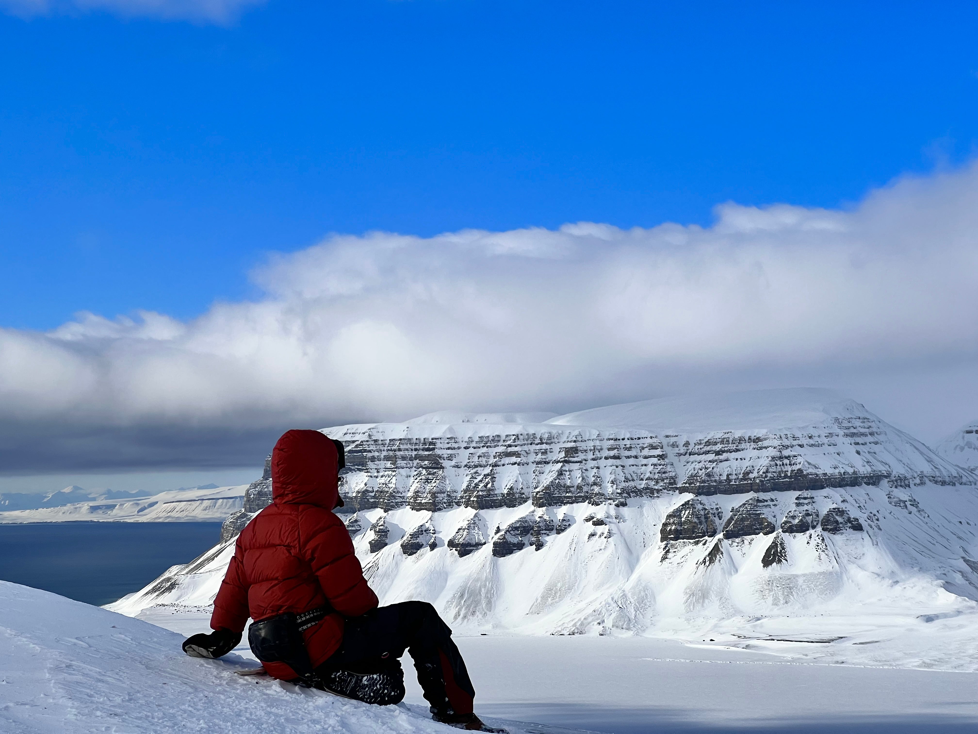 Person in a red down coat sitting on a snowy landscape with mountains and blue sky.