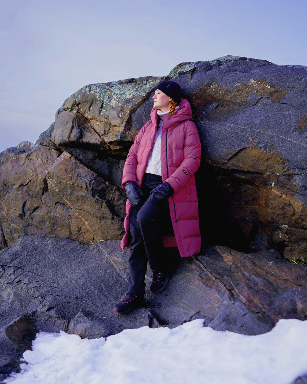A woman stands on a snowy shore in front of a large rock, wearing a long fuchsia down coat (color: peony) with a hood. Her outfit also includes dark pants, black shoes, black leather gloves, and a dark beanie.