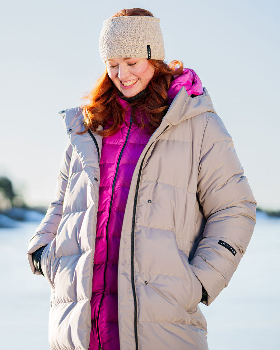 A smiling woman is outdoors in a snowy landscape wearing two layered down coats: a beige Ulrika down coat (color: cappuccino) on top and a fuchsia lightweight down jacket underneath. Her outfit also includes a light-colored knitted headband and black gloves.
