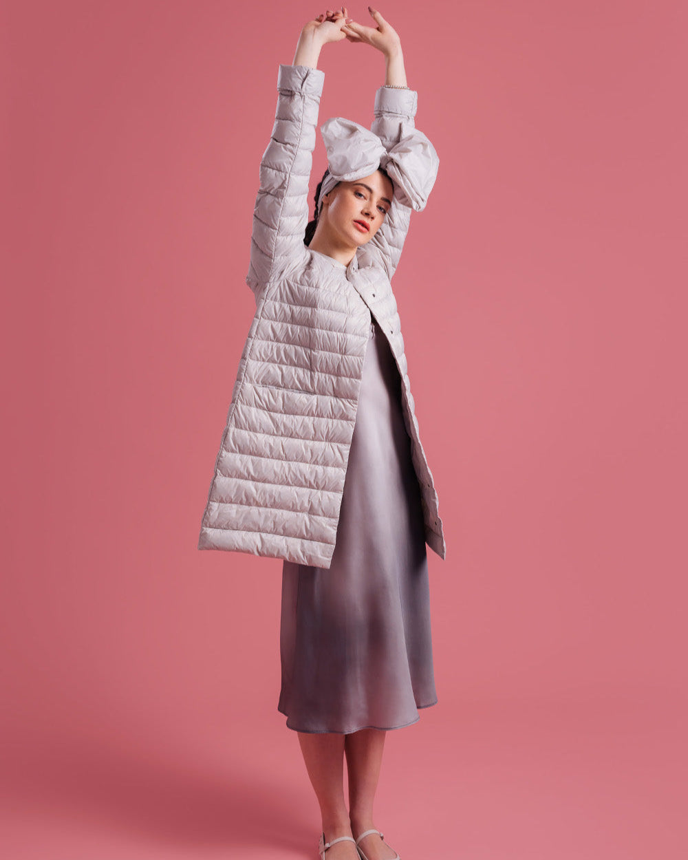 A woman stands against a pink background with her arms raised, wearing a light-colored, knee-length Vuokko light down jacket (color: sugar cloud), a shimmering gradient dress, and light-colored shoes. The outfit is completed with a large bow headband.