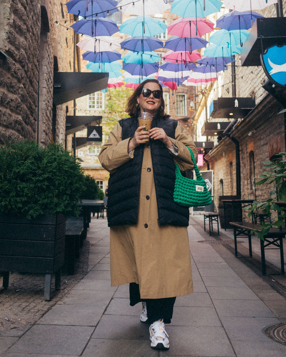 A woman is walking on a city street under colorful hanging umbrellas, wearing a black Mesi down vest (color: charcoal black) over a long beige coat. She is carrying a green bag and a takeaway drink cup.