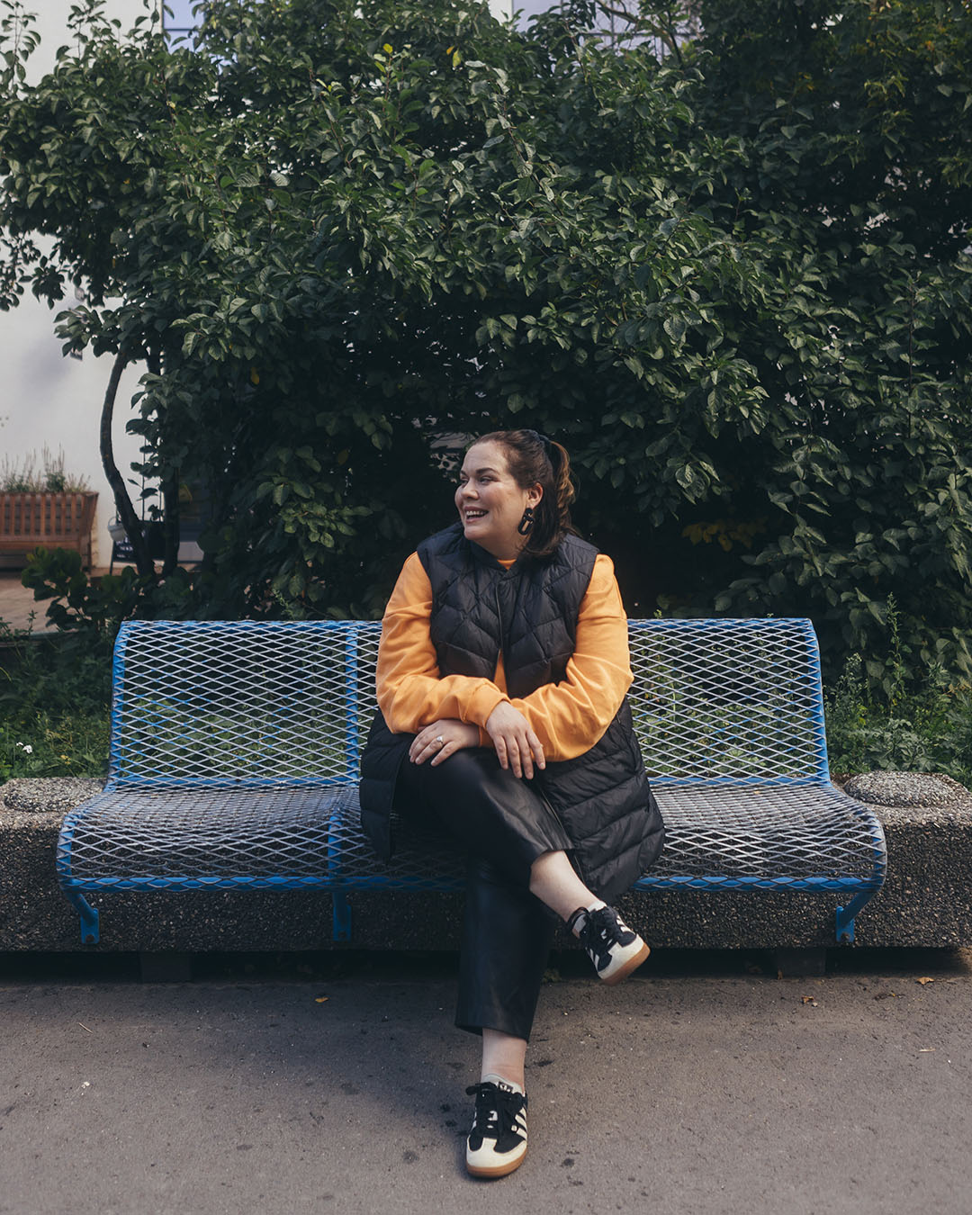 A woman is sitting outside on a blue metal bench, wearing a black Mesi down vest (color: charcoal black) and an orange shirt. She is smiling and looking to the side with her legs crossed.