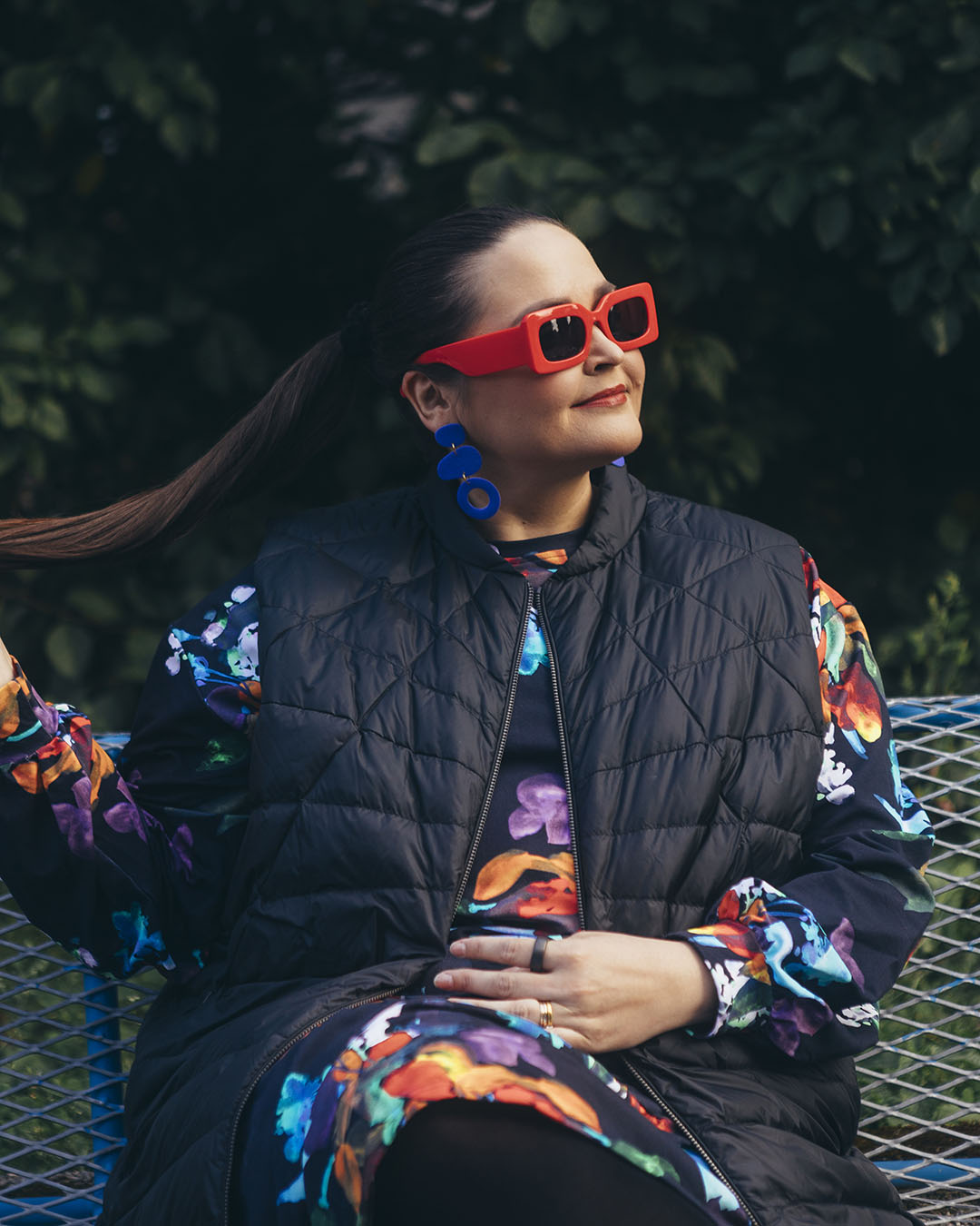 A woman is sitting on a bench wearing a black Mesi down vest (color: charcoal black) and a floral dress. She has bright red sunglasses and blue earrings, and she is holding her ponytail.