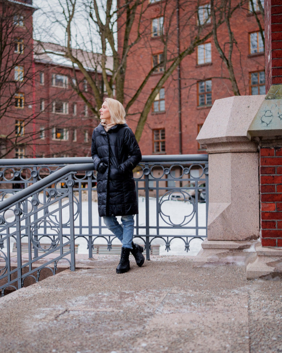 A woman stands outdoors in a winter urban environment wearing a black, long, slim-fit Essi light down coat (color: charcoal black) with a hood. Her outfit also includes blue jeans and black lace-up shoes. Brick buildings and snow-covered ground are visible in the background.