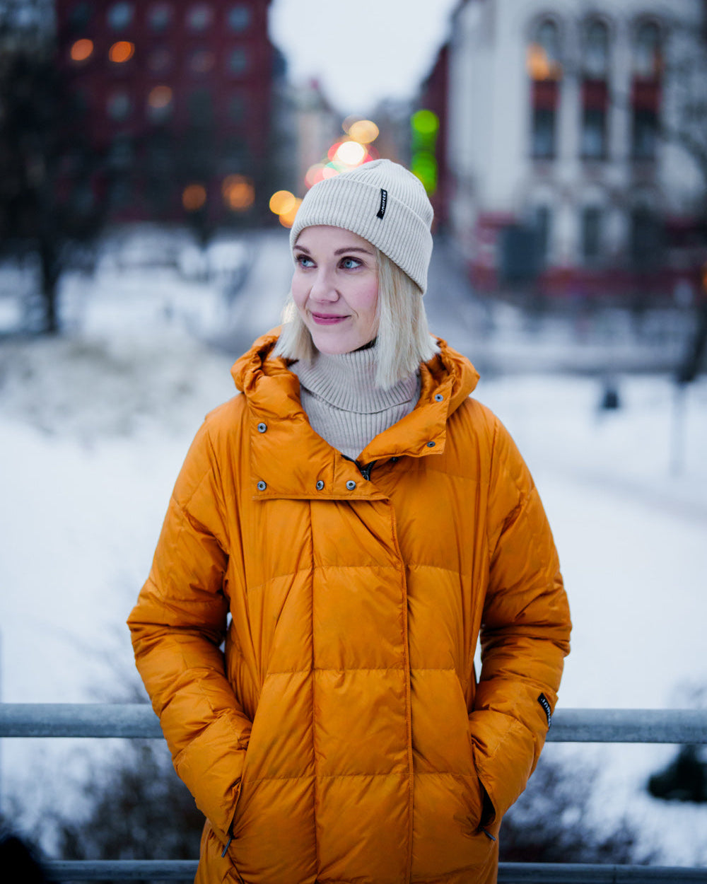 A woman stands in a winter urban setting wearing a yellow, long Essi light down coat (color: Amber) with a hood and square quilting. Her outfit also includes a light beanie and a turtleneck sweater. In the background, a snowy landscape and buildings are visible.
