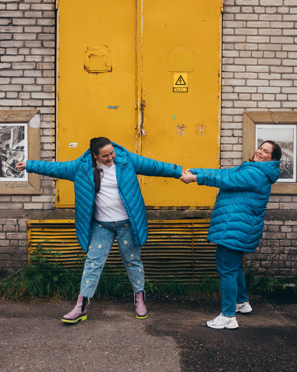 Two women are smiling in front of a yellow wall wearing blue (color: Blue butterfly) Jasmin light down coats. They are wearing blue jeans.