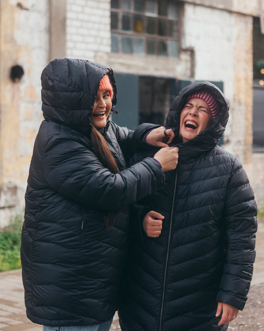 Two women are standing outside, laughing and looking at each other. Both are wearing black (color: charcoal black) quilted Jasmin light down coats with large hoods. The woman on the left has her hood up and is touching the zipper of the other woman's coat, while the woman on the right is laughing broadly with her hands down. They are both wearing knit beanies, and in the background, there's a brick wall and windows of an old building.