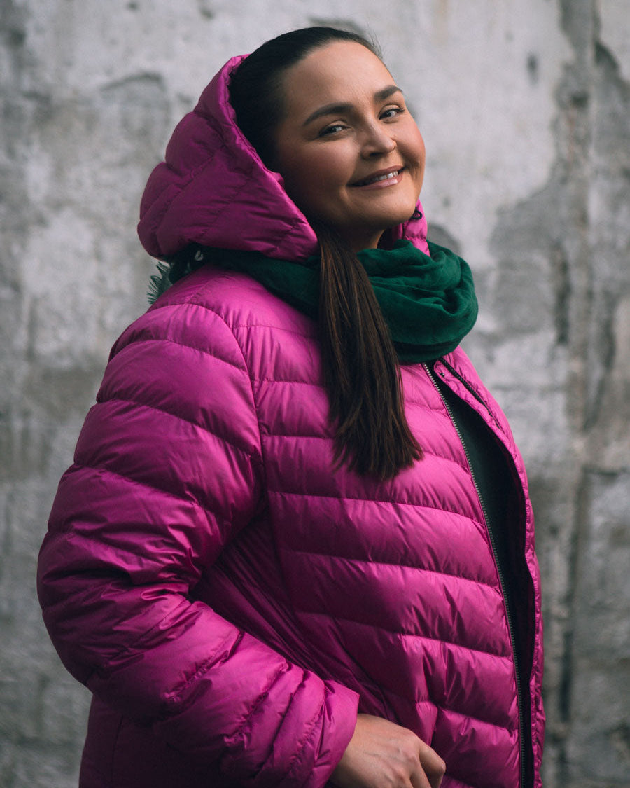 A woman is standing in front of a grey stone wall, smiling at the camera. She is wearing a bright pink (color: pitaya) Jasmin light down coat with a hood and diagonal quilting. The coat has a slightly shiny finish, and underneath she is wearing a green scarf that adds a splash of color to the outfit.