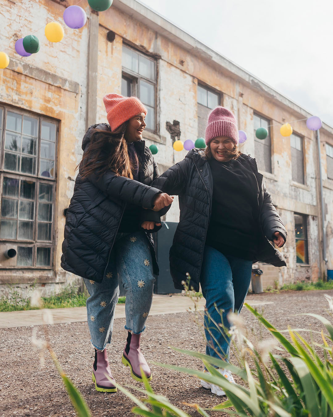 Two women are laughing and walking hand in hand outside in front of an old building. Both are wearing charcoal black Jasmin light down coats and colorful beanies.