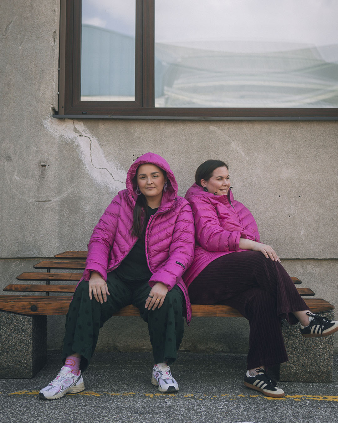 Two women are sitting outside side by side on a bench, wearing fuchsia Jasmin light down coats (color: pitaya). The coats are quilted and hooded. In the background, there is a grey plastered wall and a large window.